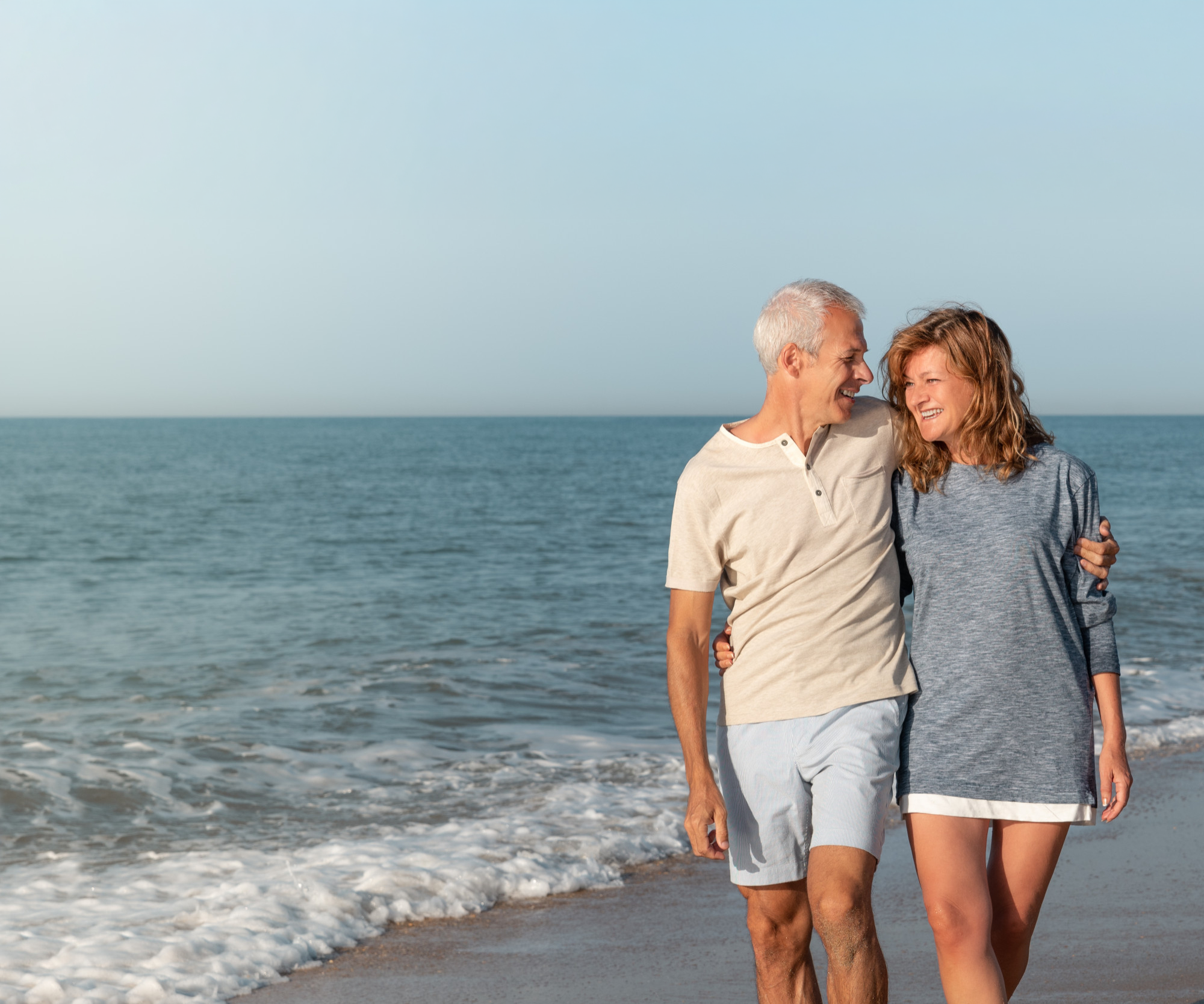 An elderly couple walking along the beach, smiling and holding hands with the ocean and sky in the background.