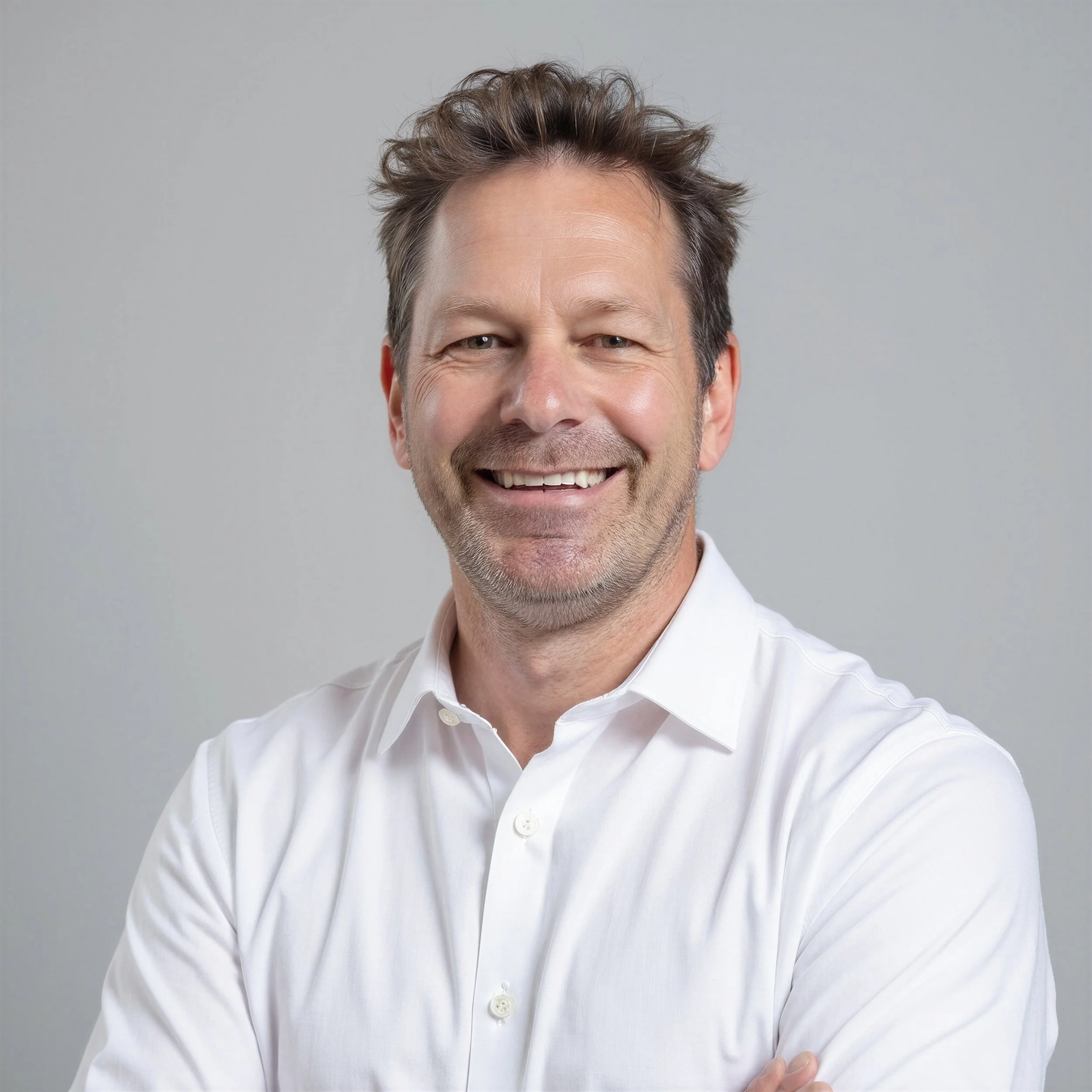 A smiling middle-aged man with short, darker hair that is graying, wearing a white dress shirt, posed against a plain gray background.