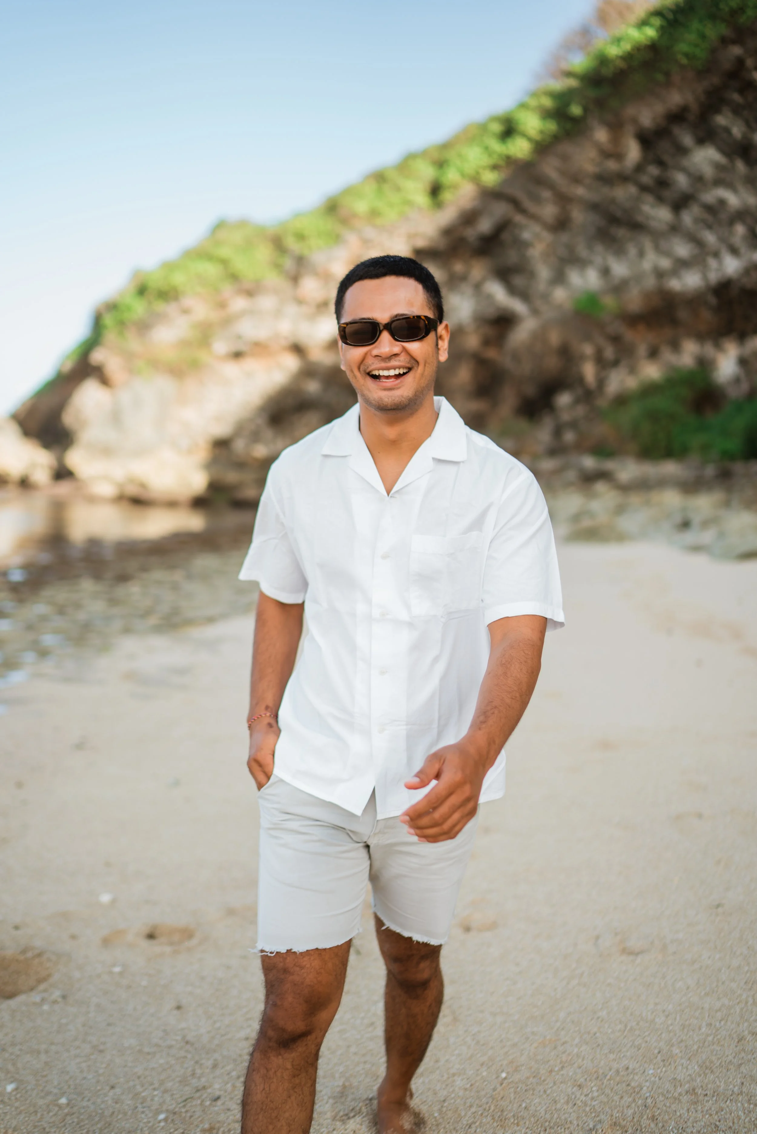 A smiling man wearing sunglasses and a white short-sleeved shirt walking on a beach with rocks and green shrubbery in the background.