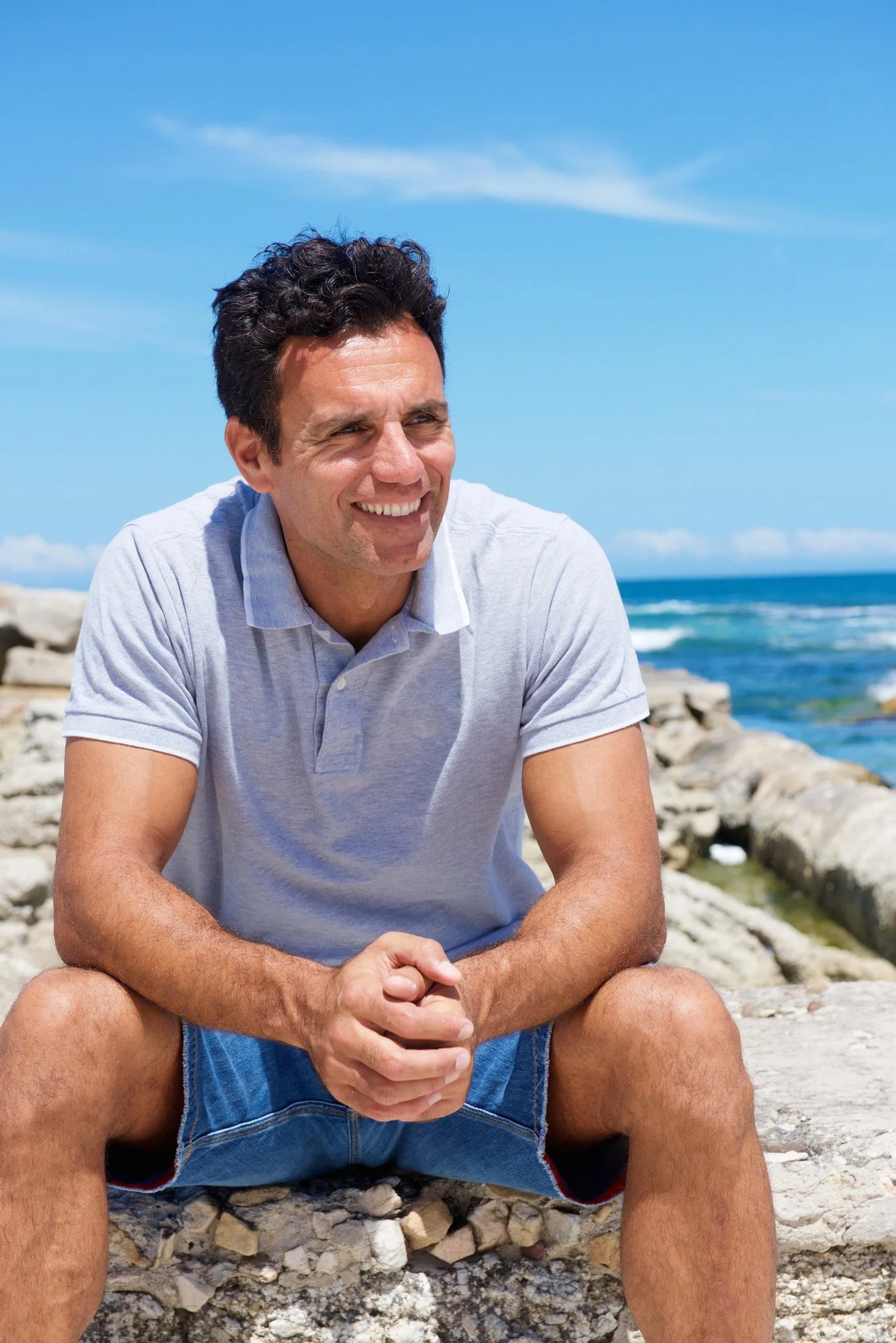 A smiling man with dark curly hair wearing a light gray polo shirt and denim shorts, sitting on rocks near the beach with the ocean and blue sky in the background.