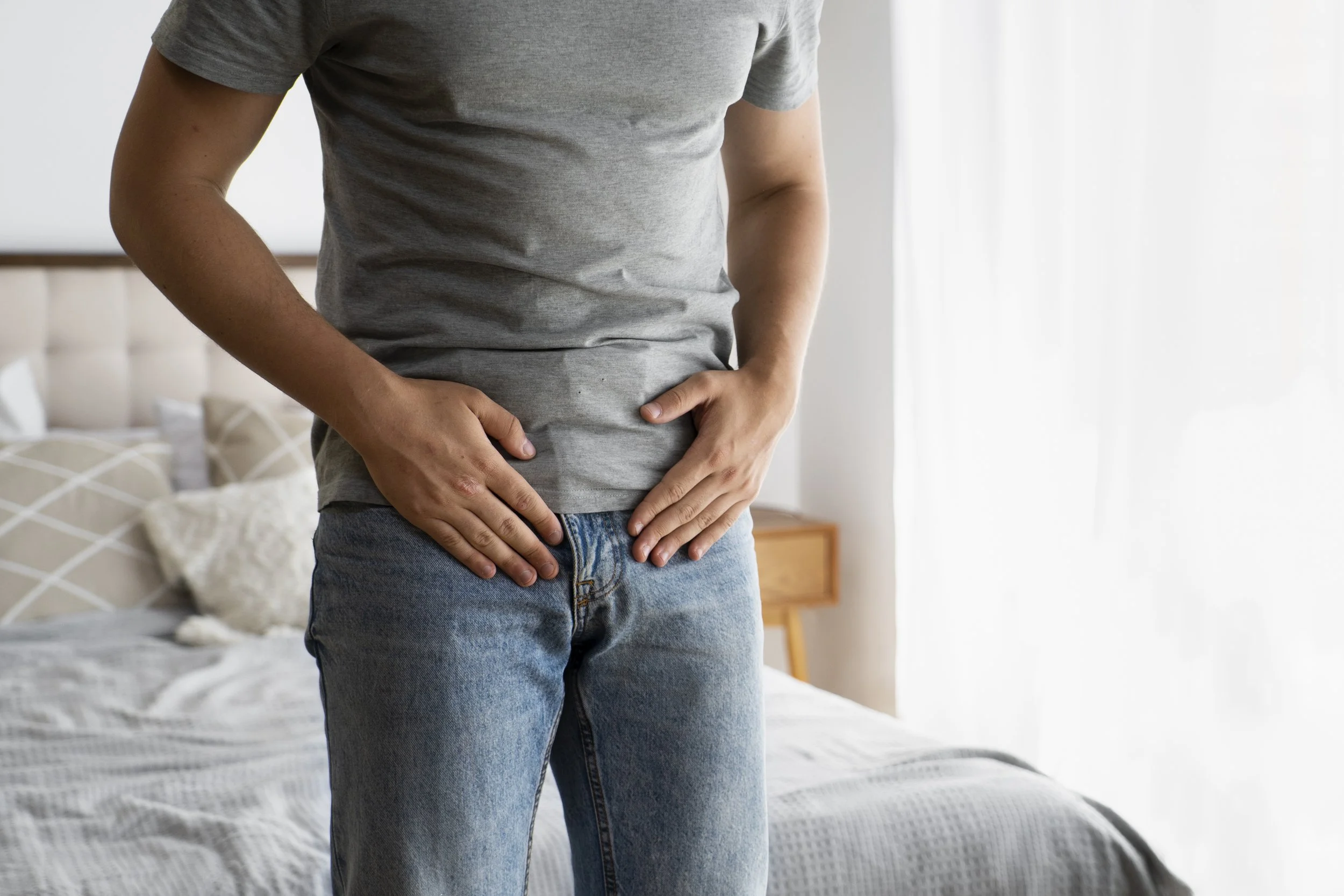 Person wearing gray t-shirt and blue jeans standing in a bedroom, with hands placed on stomach.