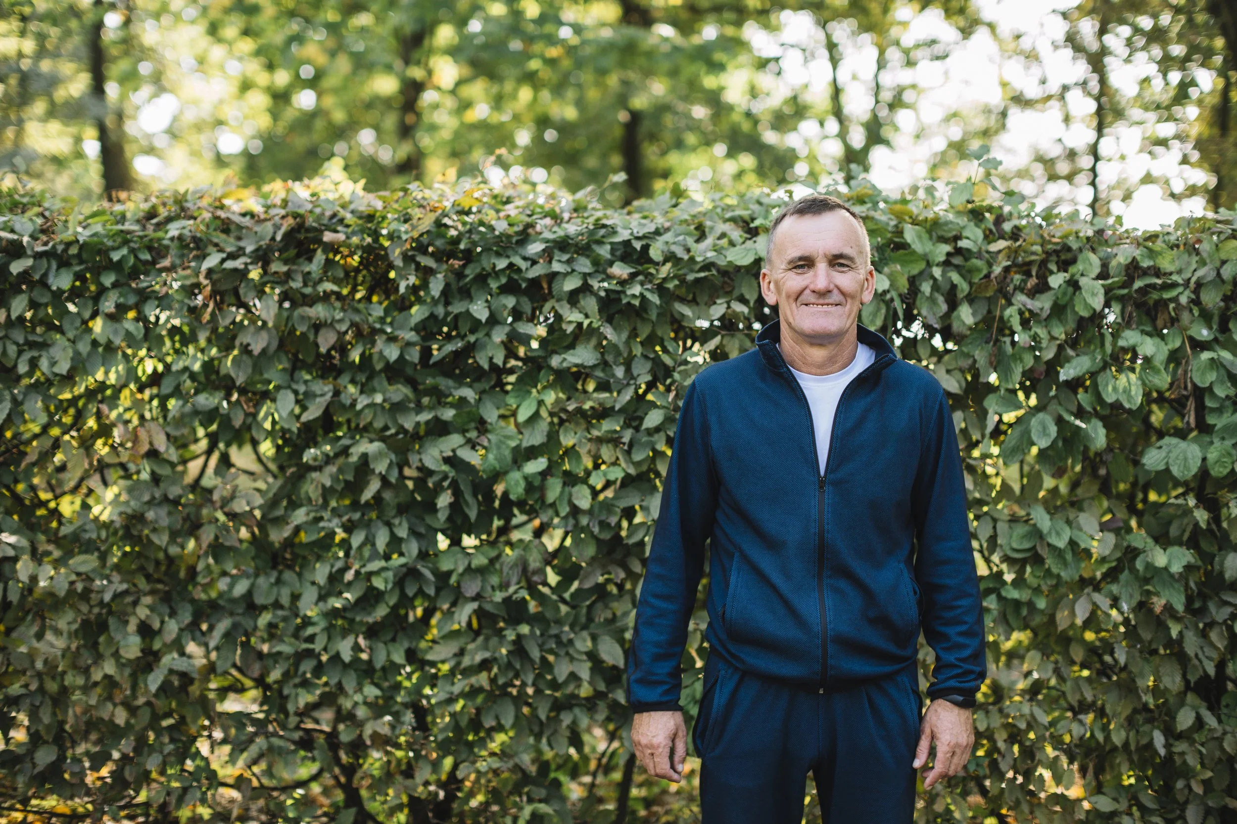 A middle-aged man standing outdoors in front of a lush, green hedge, smiling at the camera, wearing a navy blue zip-up jacket and a white shirt underneath.