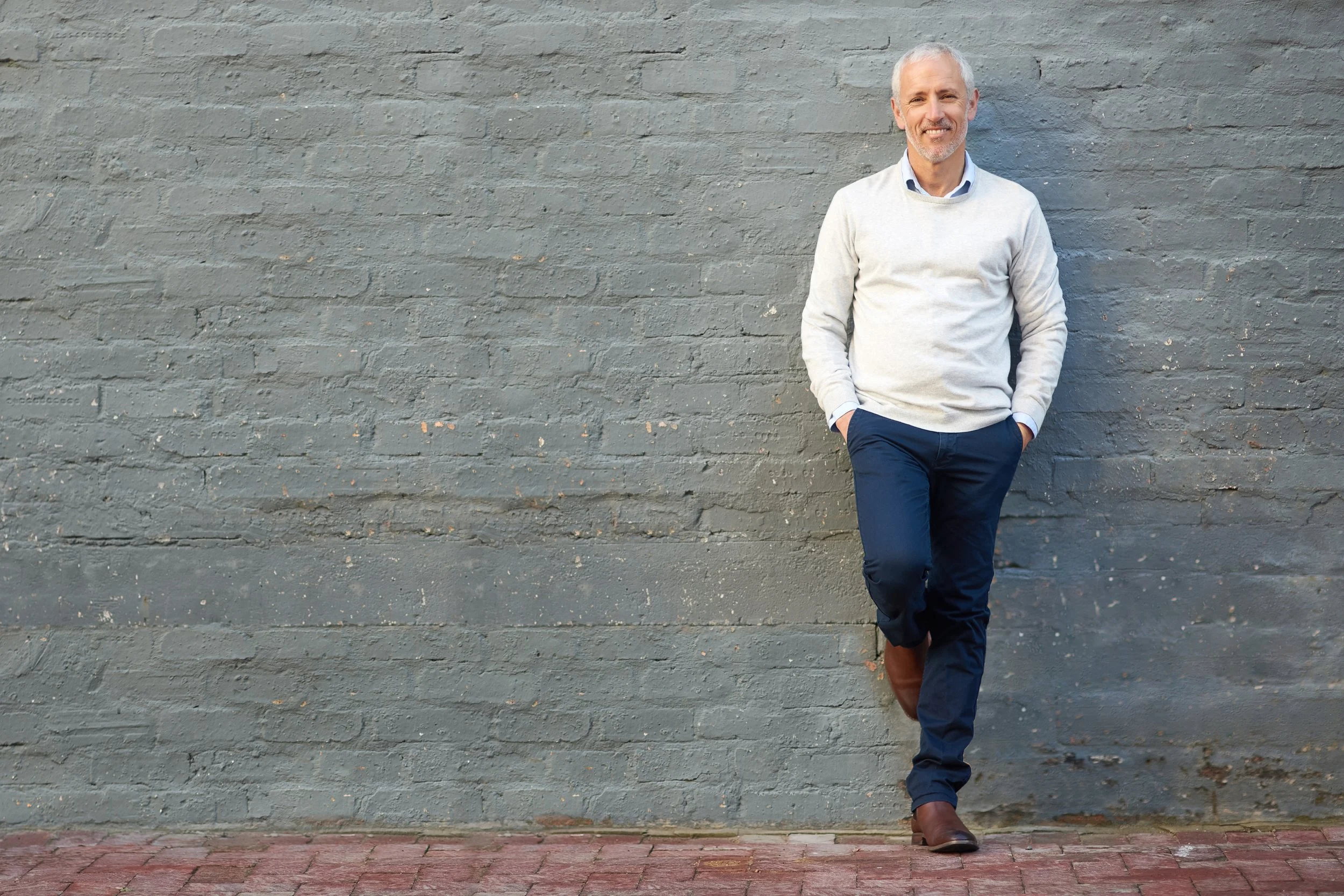 A middle-aged man with gray hair and a beard, dressed in a white sweater, blue shirt, and dark pants, leaning casually against a gray brick wall, smiling.