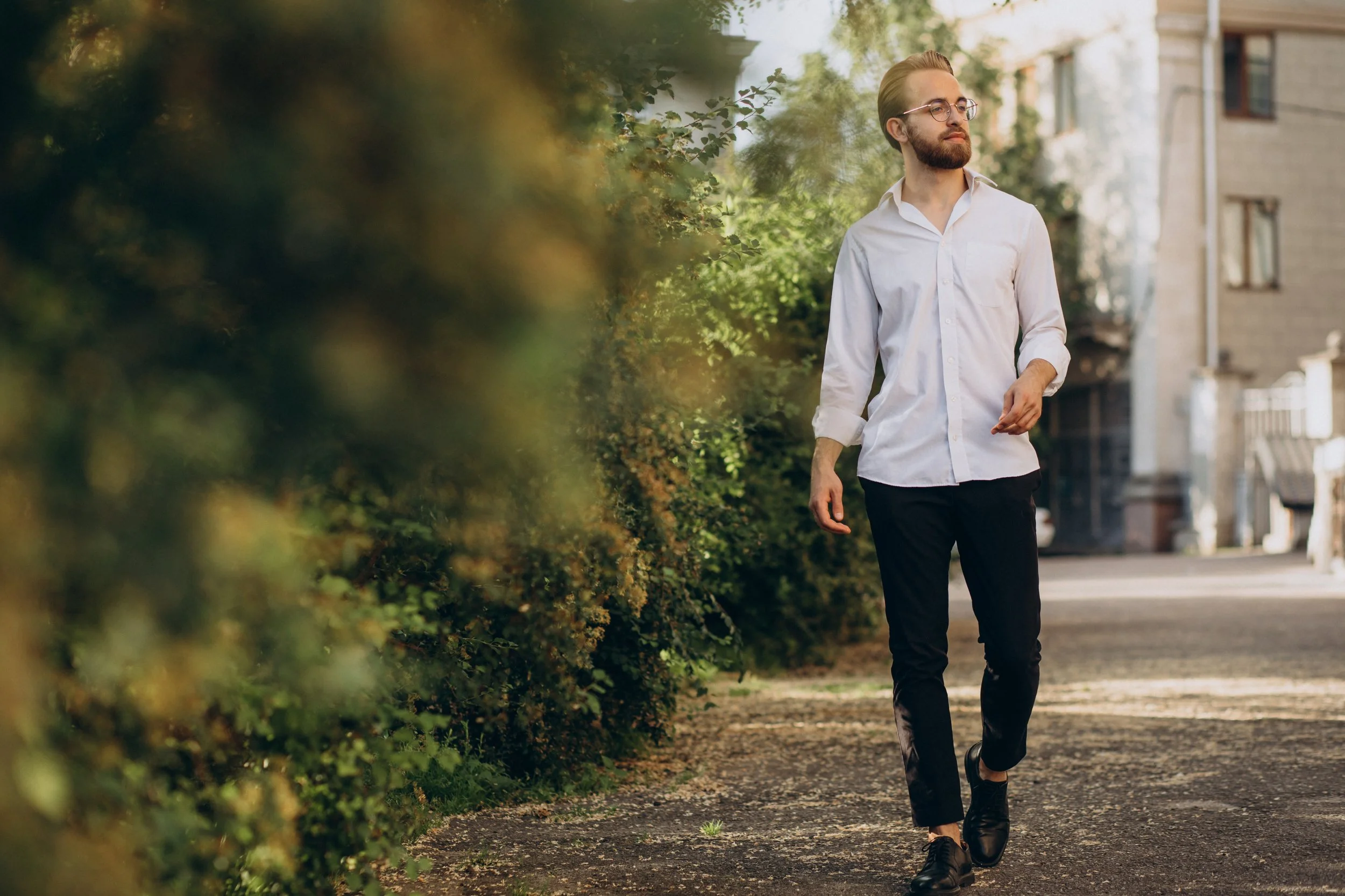 A man with glasses, a beard, and dressed in a white shirt and black pants, is walking along a sidewalk bordered by greenery and residential buildings on a sunny day.