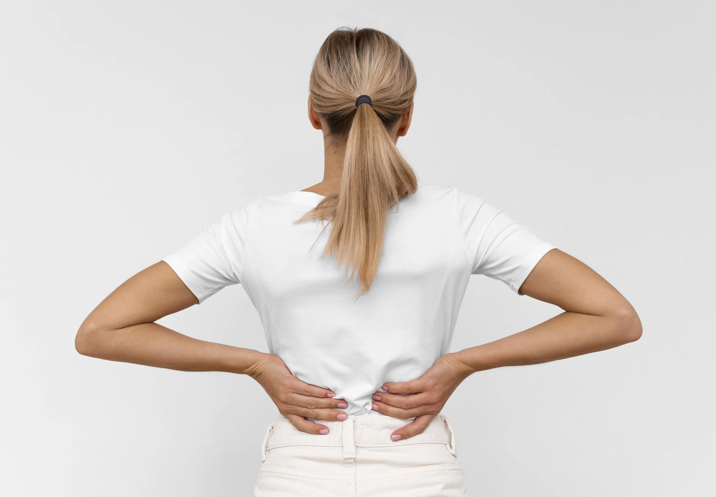 Back view of woman with blonde hair tied in a ponytail, wearing a white shirt, holding her lower back with both hands, suggesting she might be experiencing discomfort.