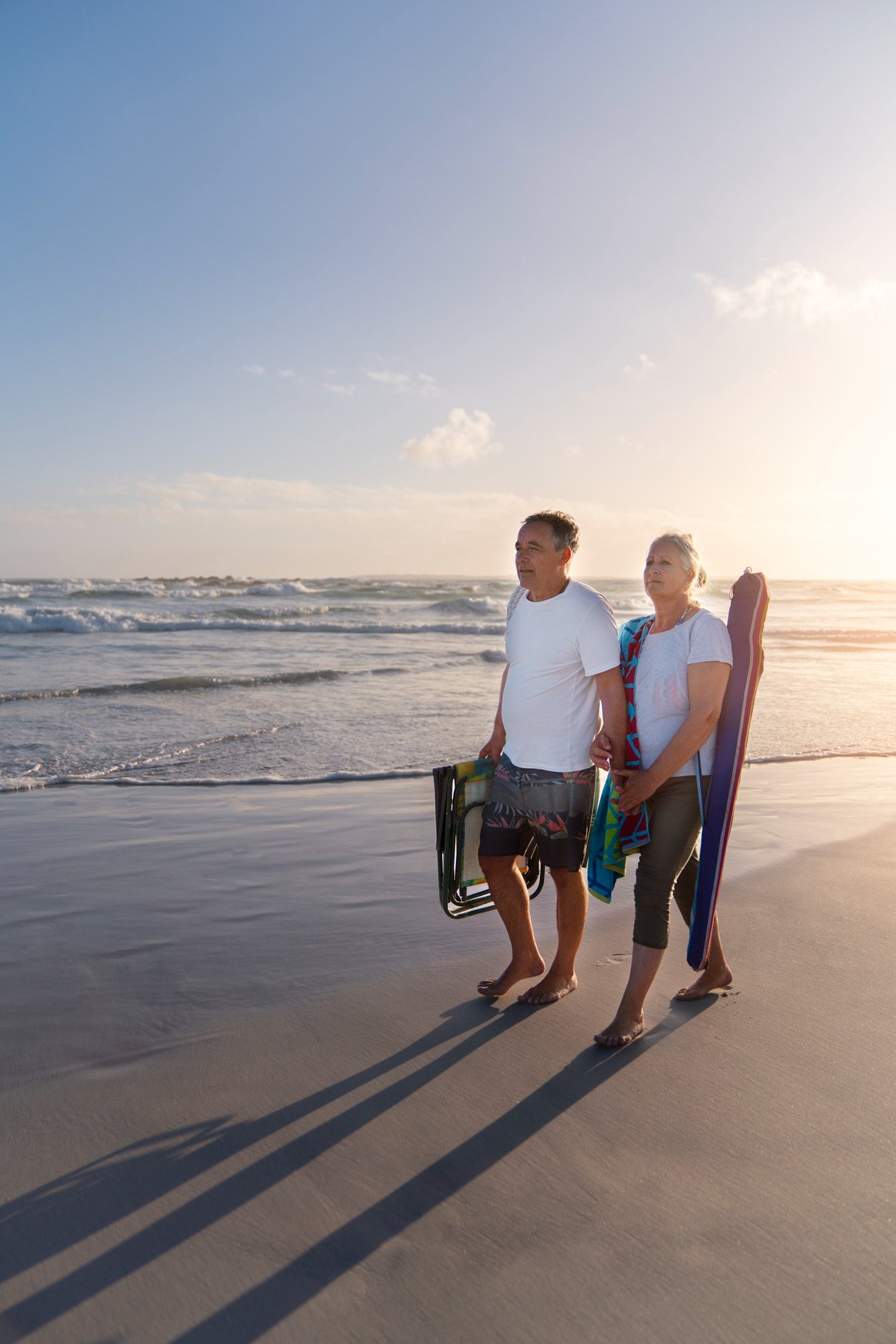 An elderly couple walking barefoot on the beach with their shadow cast on the sand during sunset. The man is pushing a lawn chair, and the woman is carrying a surfboard; both are dressed casually.