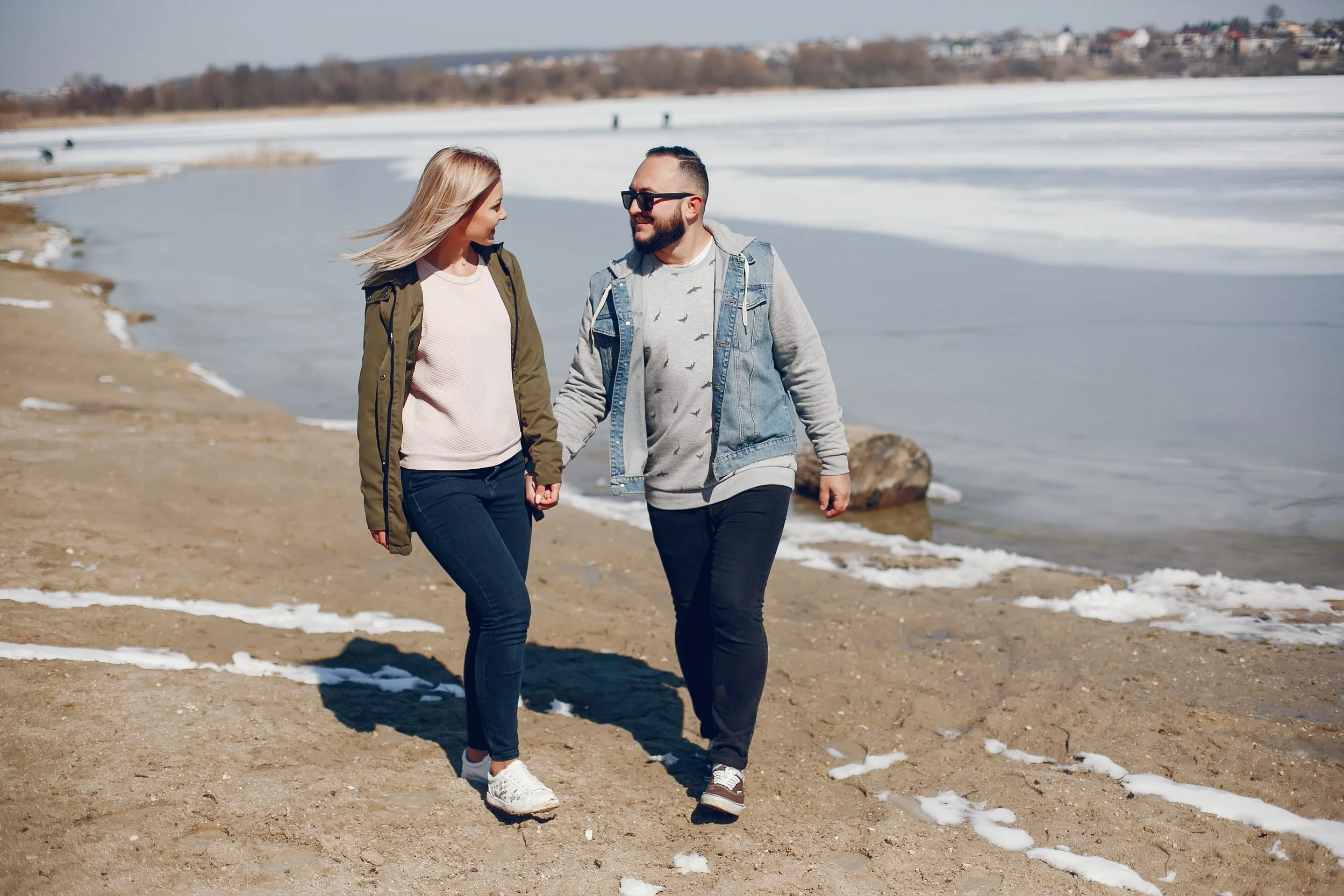 A young couple holding hands and walking on a sandy beach near a body of water with snow and ice, in a cold weather setting.