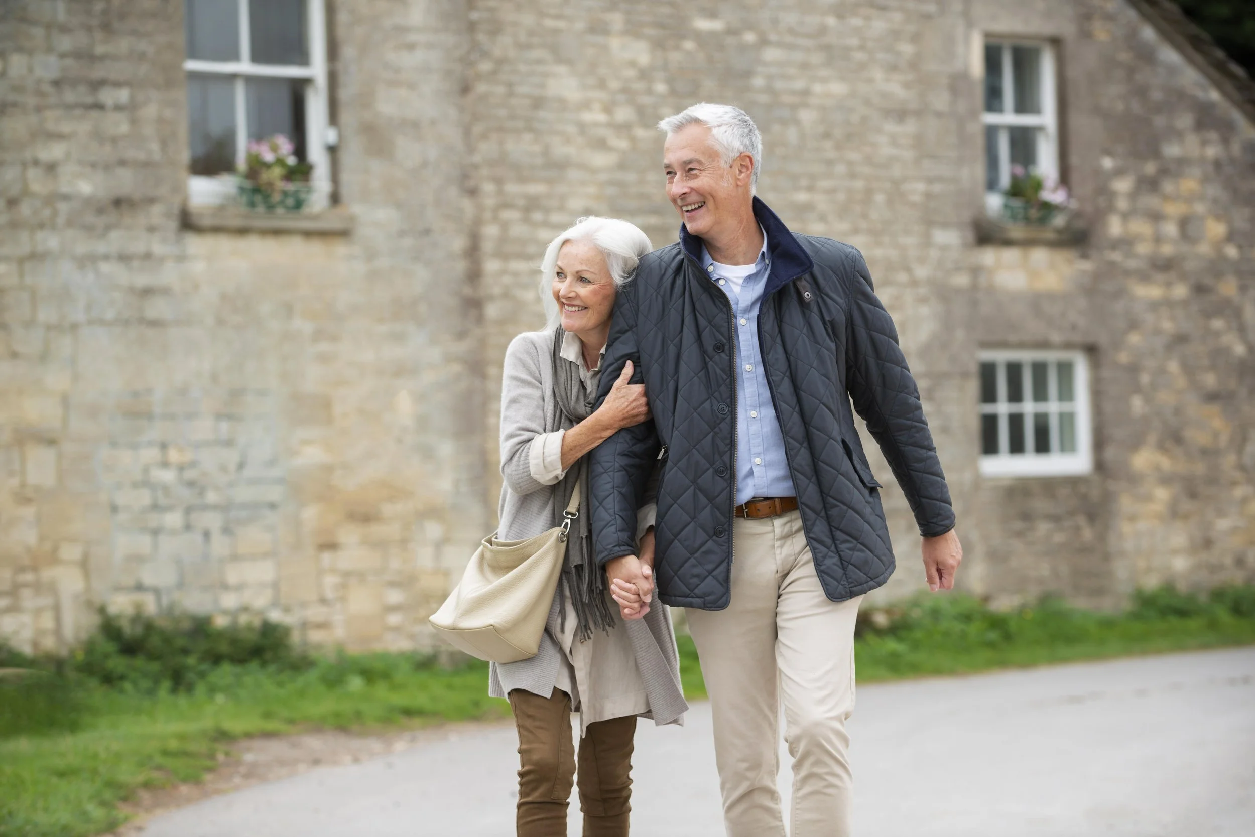 An elderly couple holding hands and smiling while walking outdoors near a brick building with windows and flower pots, dressed casually in jackets and beige pants.