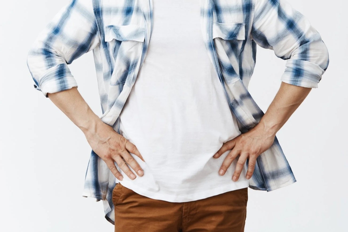 Person wearing a white t-shirt and a plaid shirt with rolled-up sleeves, hands on hips, against a plain background.