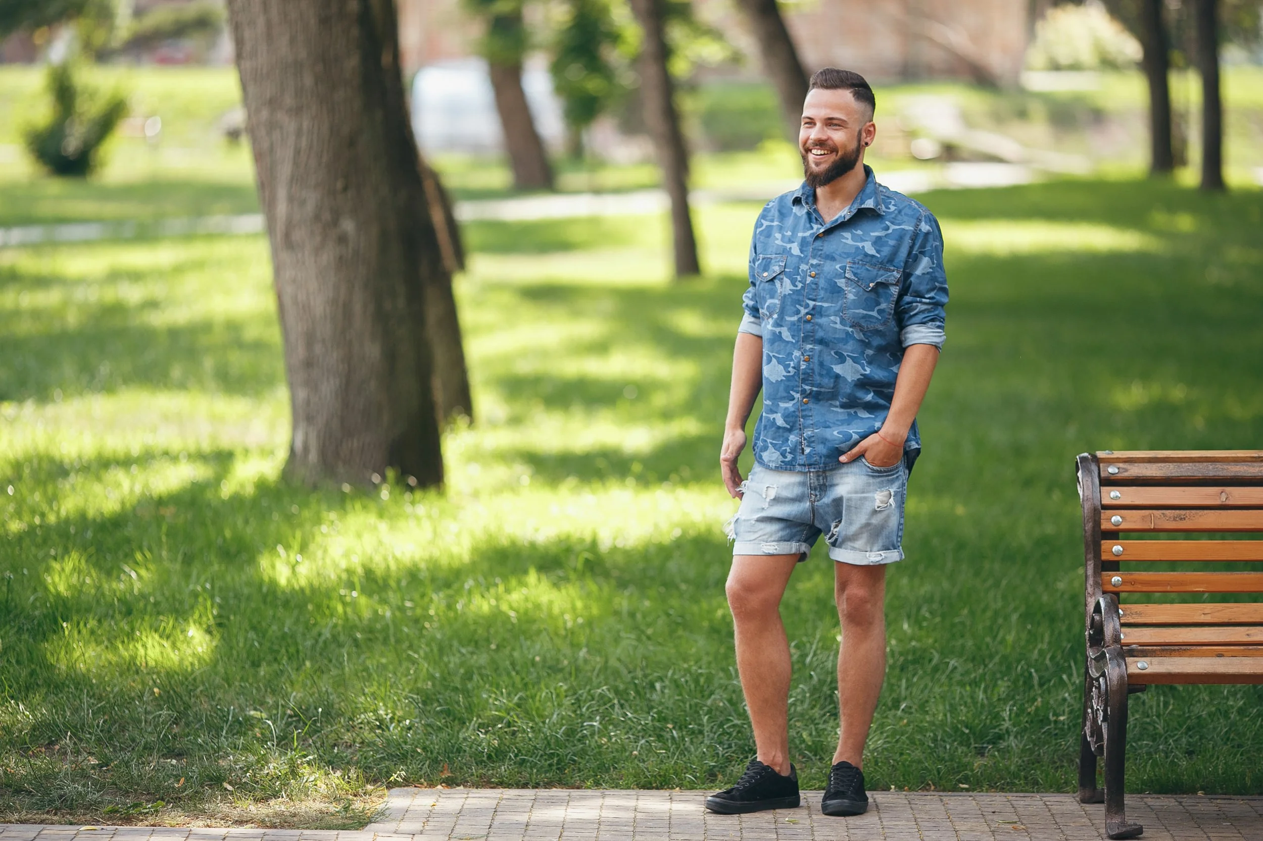 A young man with a beard and short hair, wearing a blue camouflage shirt and ripped denim shorts, standing outdoors in a park with green grass and trees, smiling and looking to his left near a wooden park bench.
