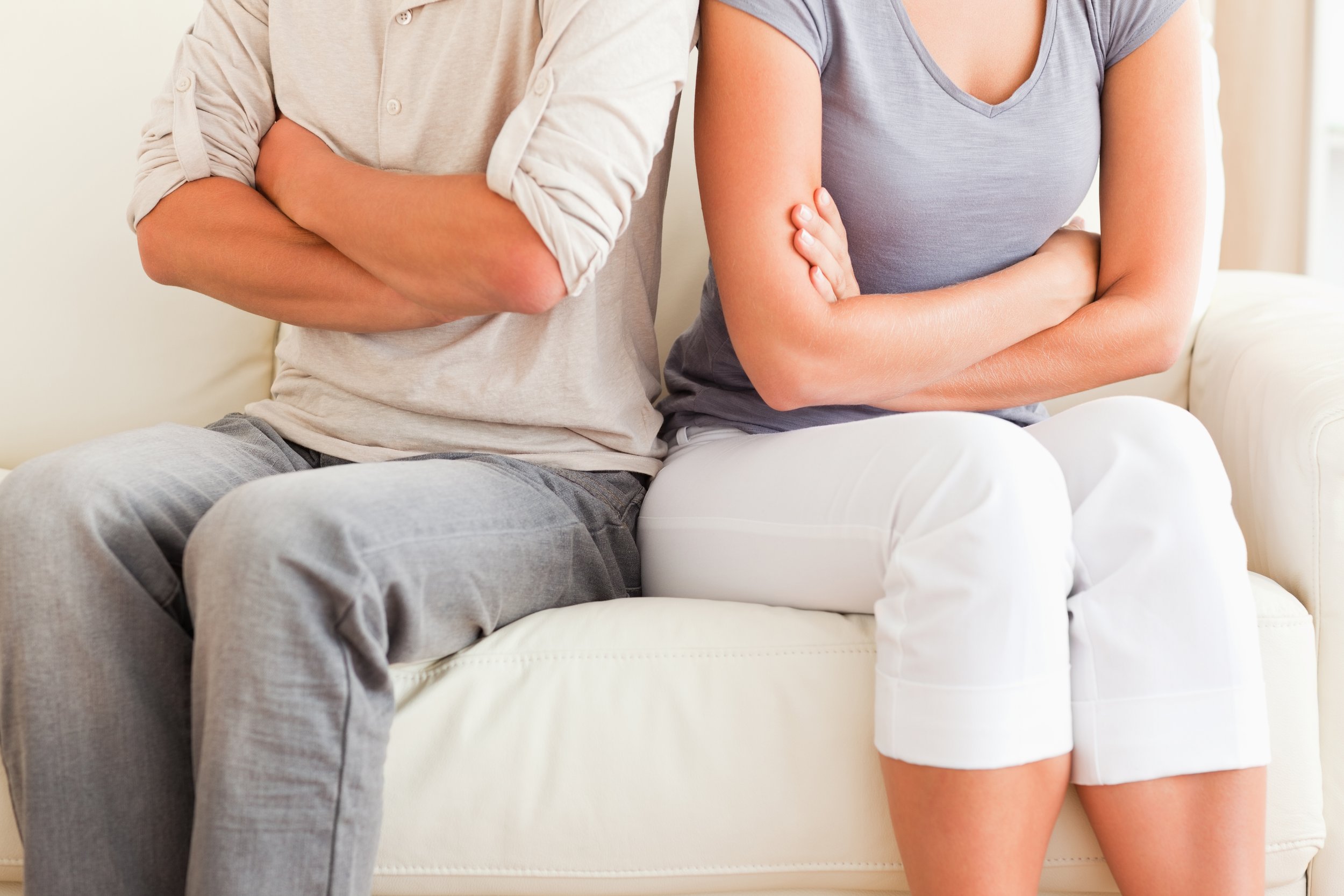 A man and a woman sitting on a cream-colored couch with their arms crossed, the man wearing a beige shirt and gray pants, and the woman wearing a gray T-shirt and white shorts.