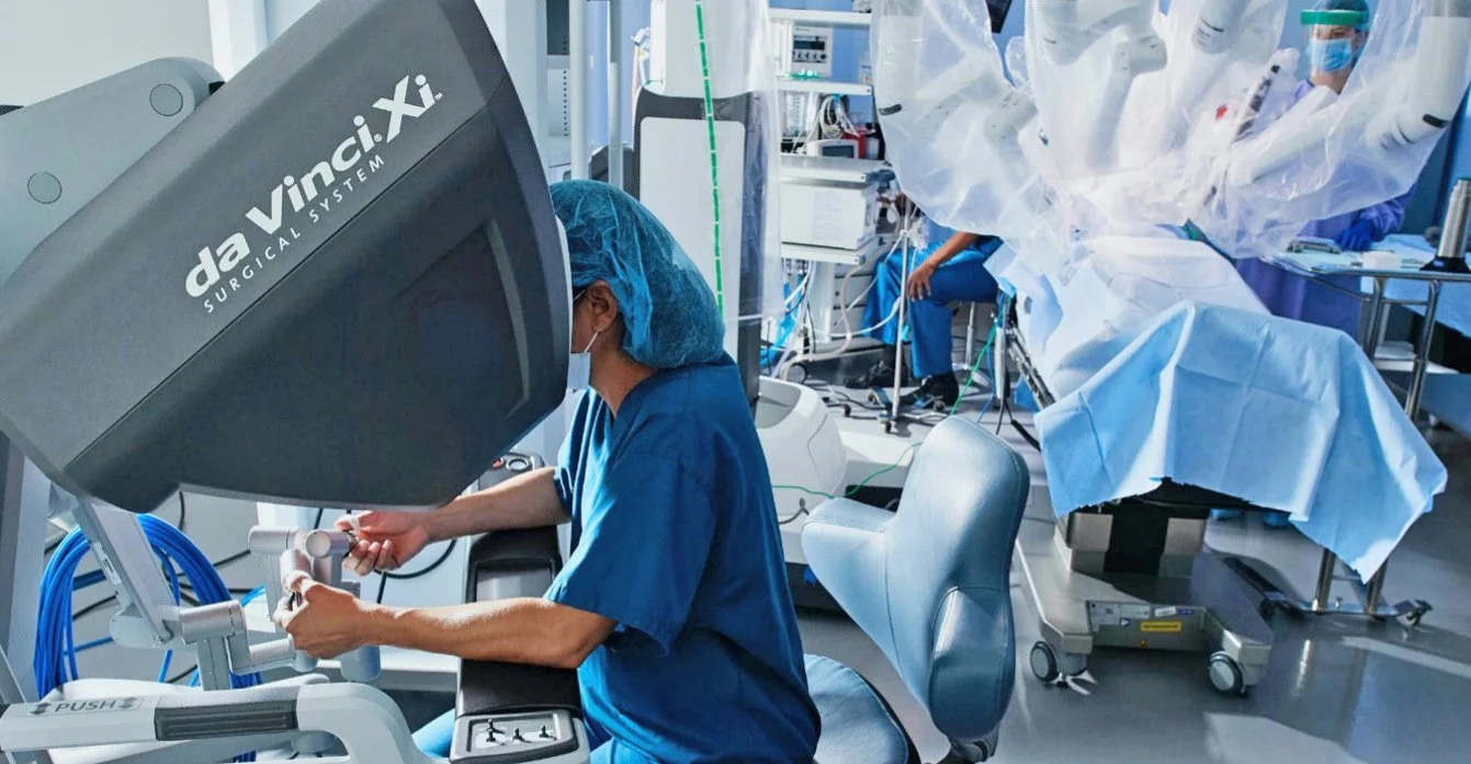 A surgeon operating in an operating room with a da Vinci Surgical System robotic arm and medical personnel in scrubs and masks.