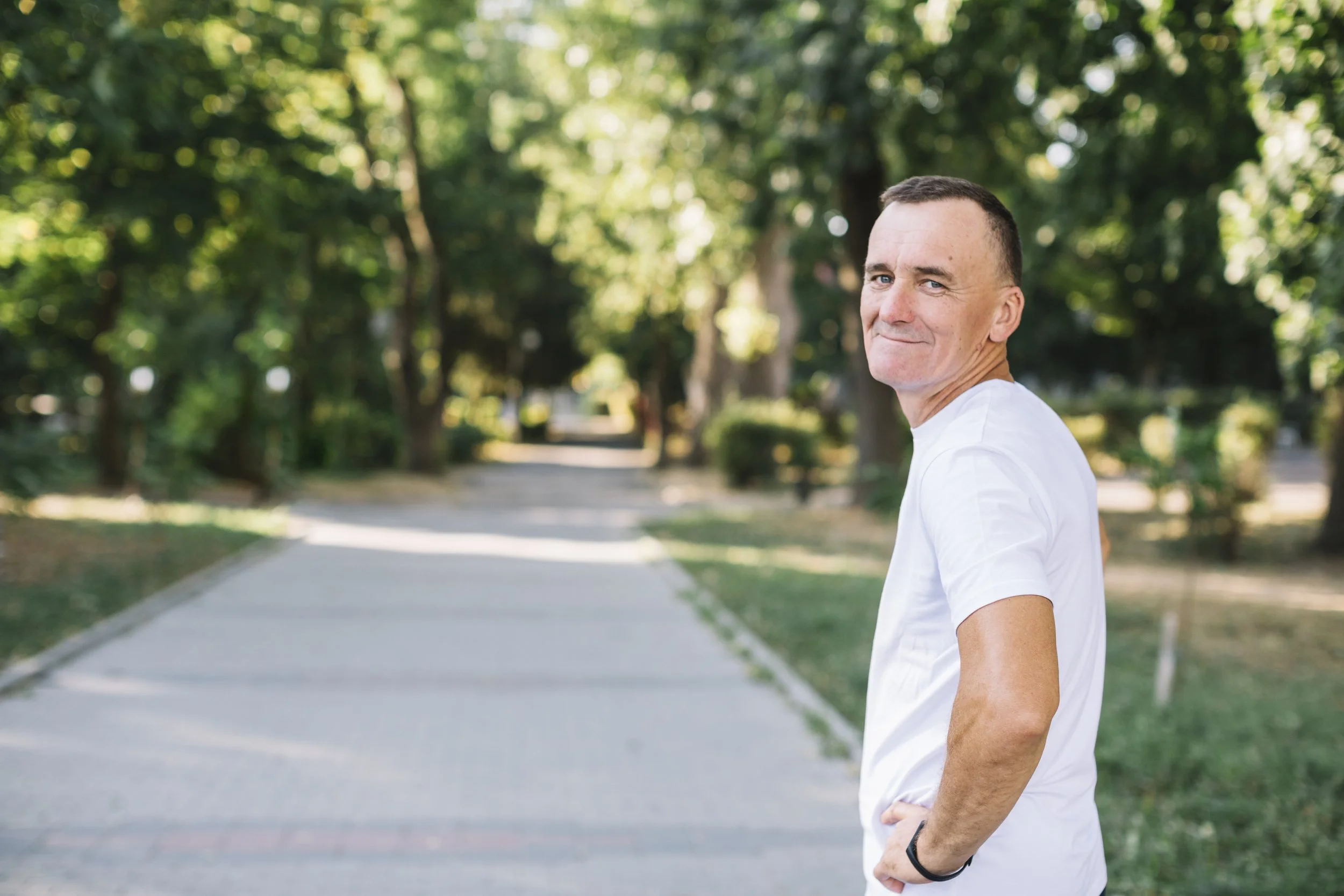 A man with short dark hair and blue eyes smiling while standing outdoors on a tree-lined path, wearing a white t-shirt.