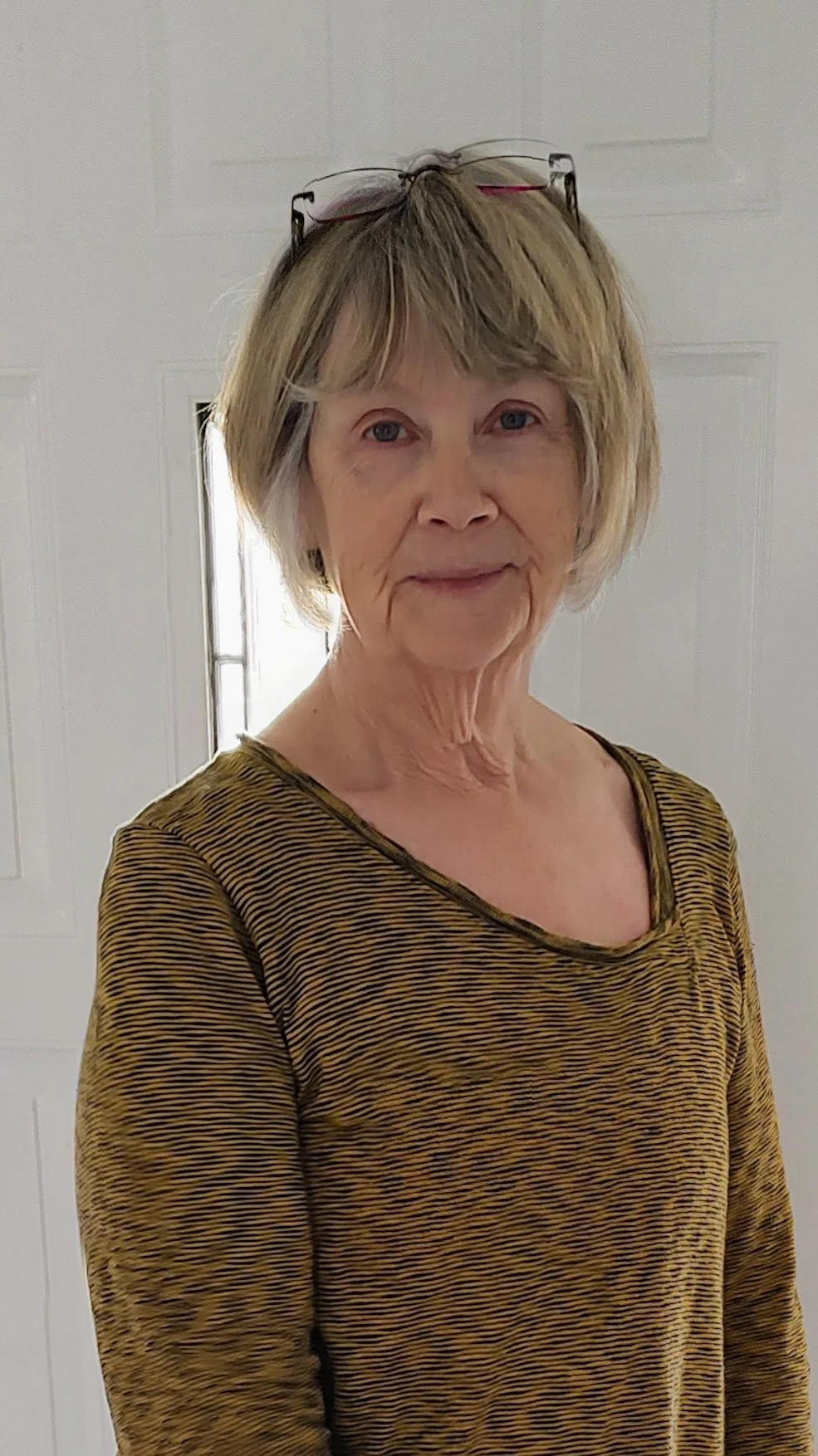 Older woman with short blonde hair wearing glasses on her head and a striped long-sleeve shirt, standing indoors in front of a white door.