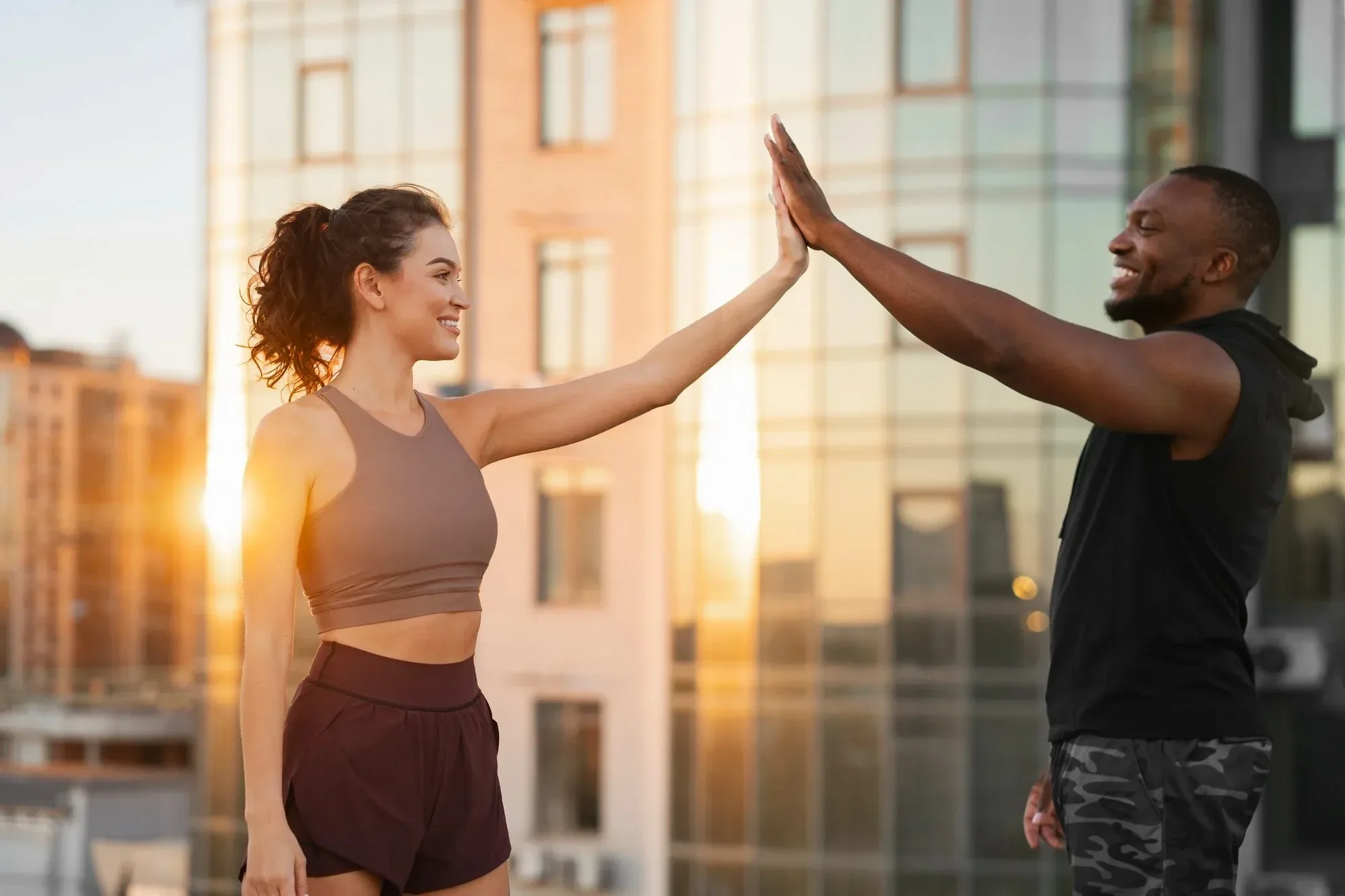 A man and woman giving each other a high five outdoors, with a cityscape background during sunset, both smiling, dressed in athletic clothing.