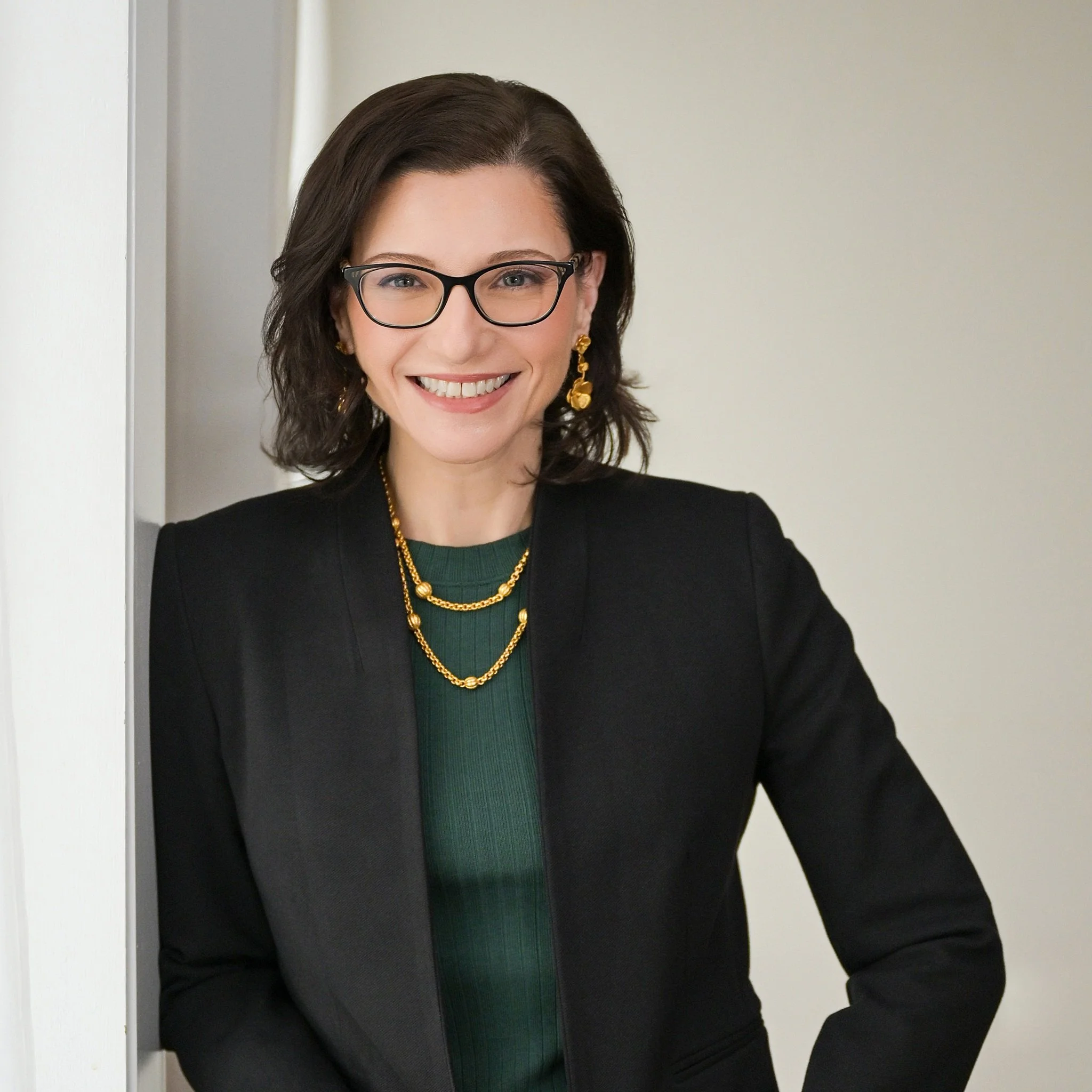 A woman with shoulder-length dark hair, glasses, and a bright smile, wearing a black blazer, green top, and gold jewelry, standing indoors against a light-colored wall.