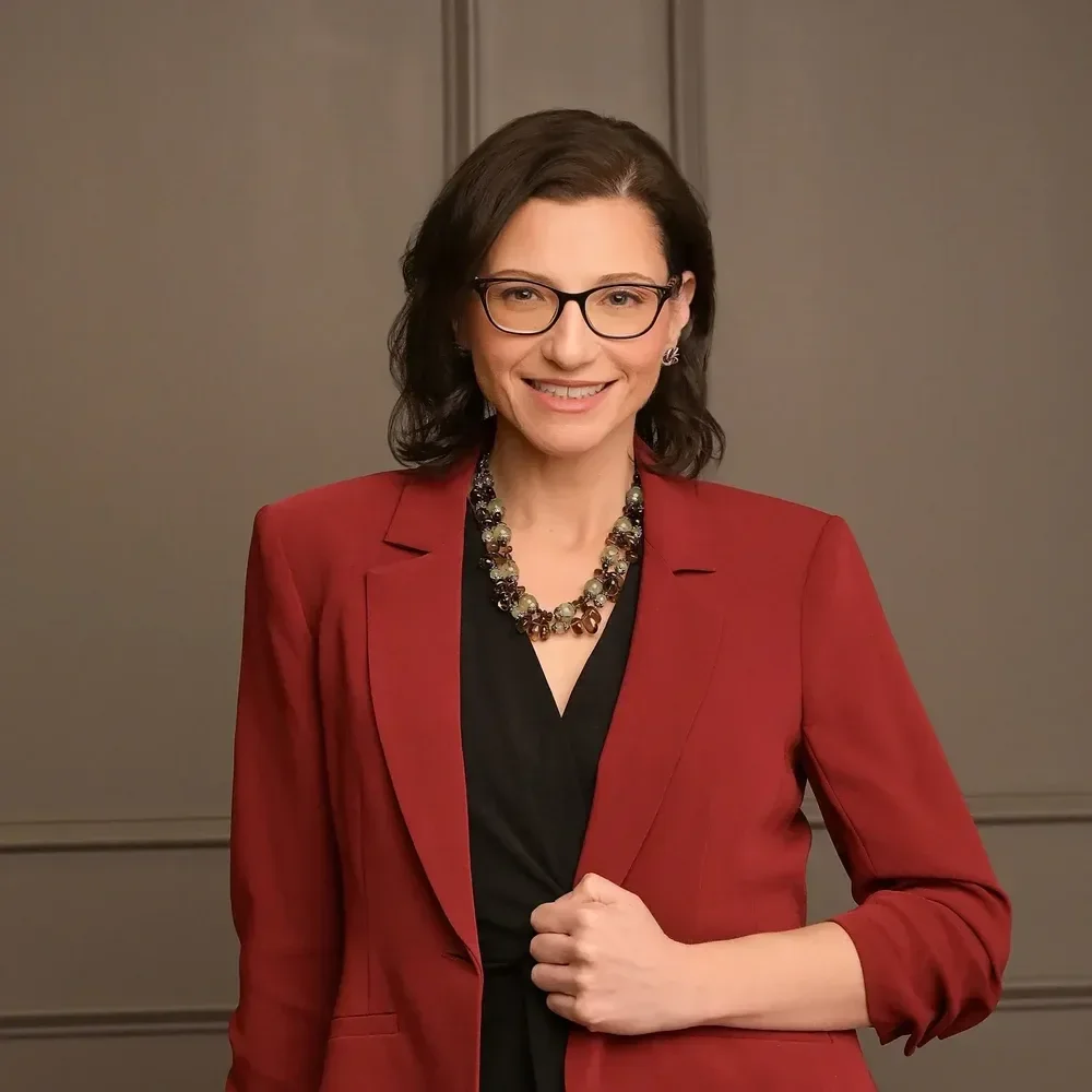 Dr. Marisa Mizus smiling with glasses, wearing a red blazer over a black top, accessorized with a beaded necklace and earrings, standing in front of a plain wall.