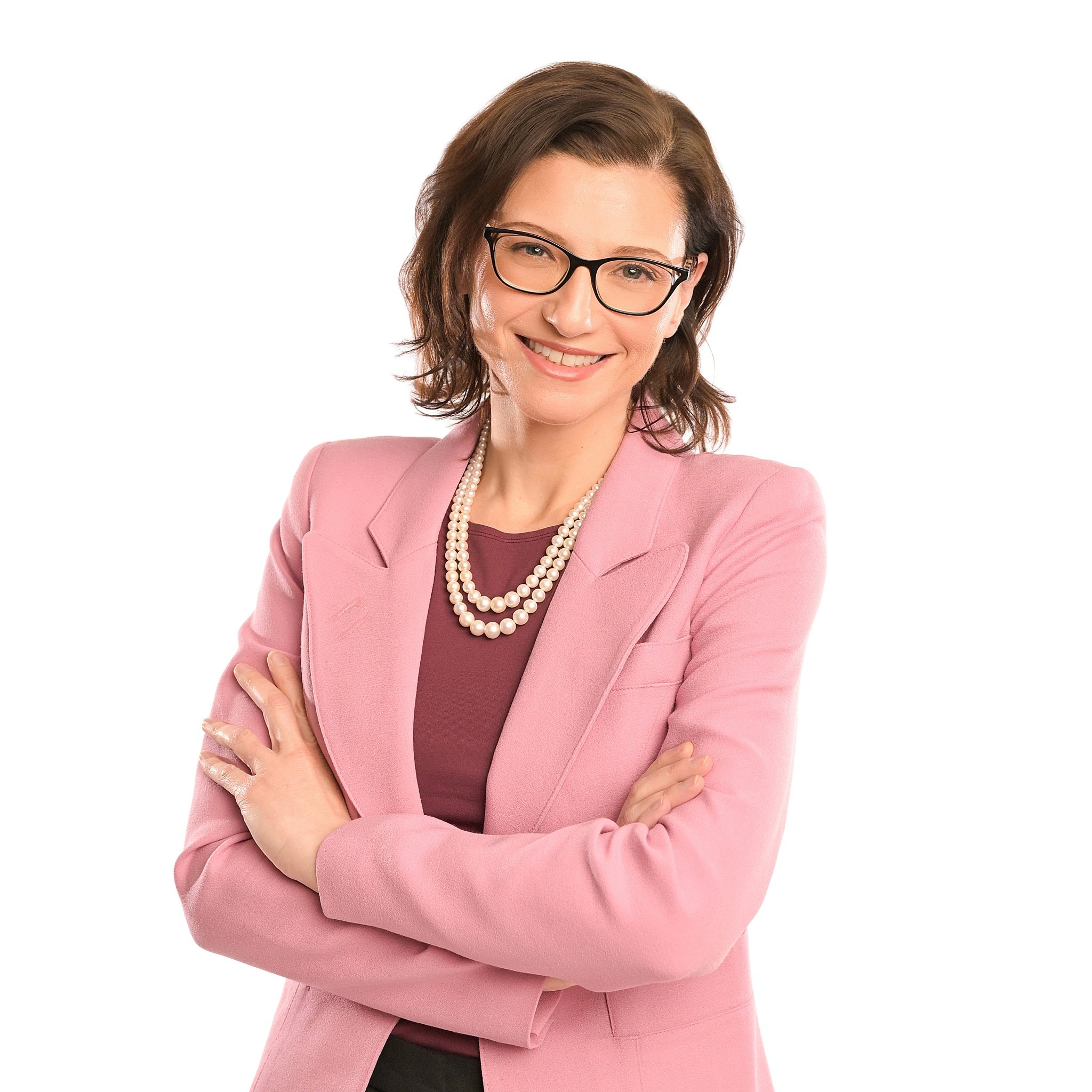A woman physician with brown hair, glasses, and a friendly smile, wearing a pink blazer, pearl necklace, and a maroon top, standing with her arms crossed against a white background.