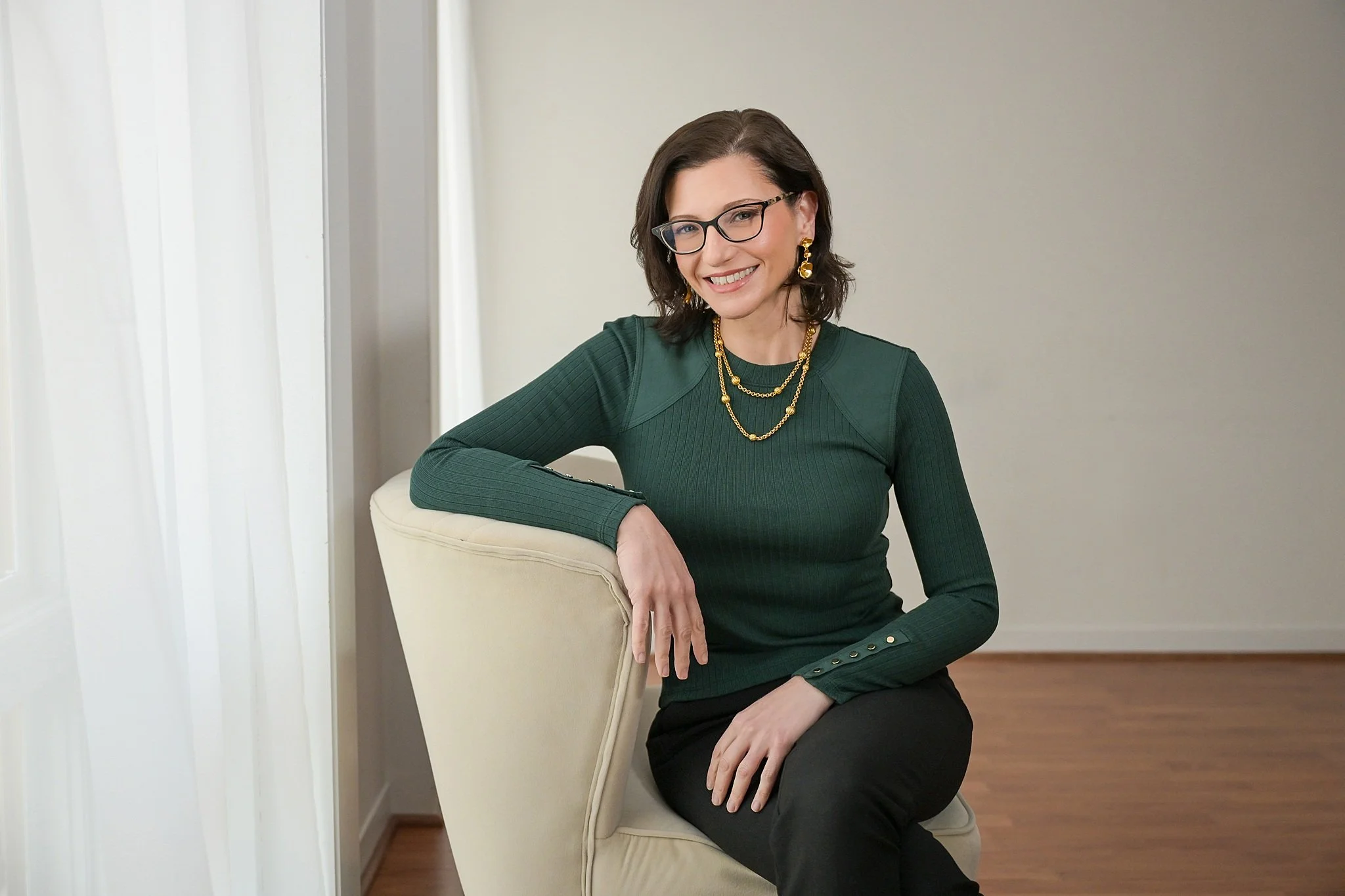 A woman physician with glasses, wearing a green sweater with button details on the sleeves, gold jewelry, and black pants, sitting on a white chair in a room with hardwood floors and neutral walls.