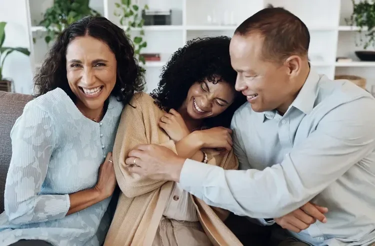 Three adults laughing and hugging each other on a couch in a bright living room.