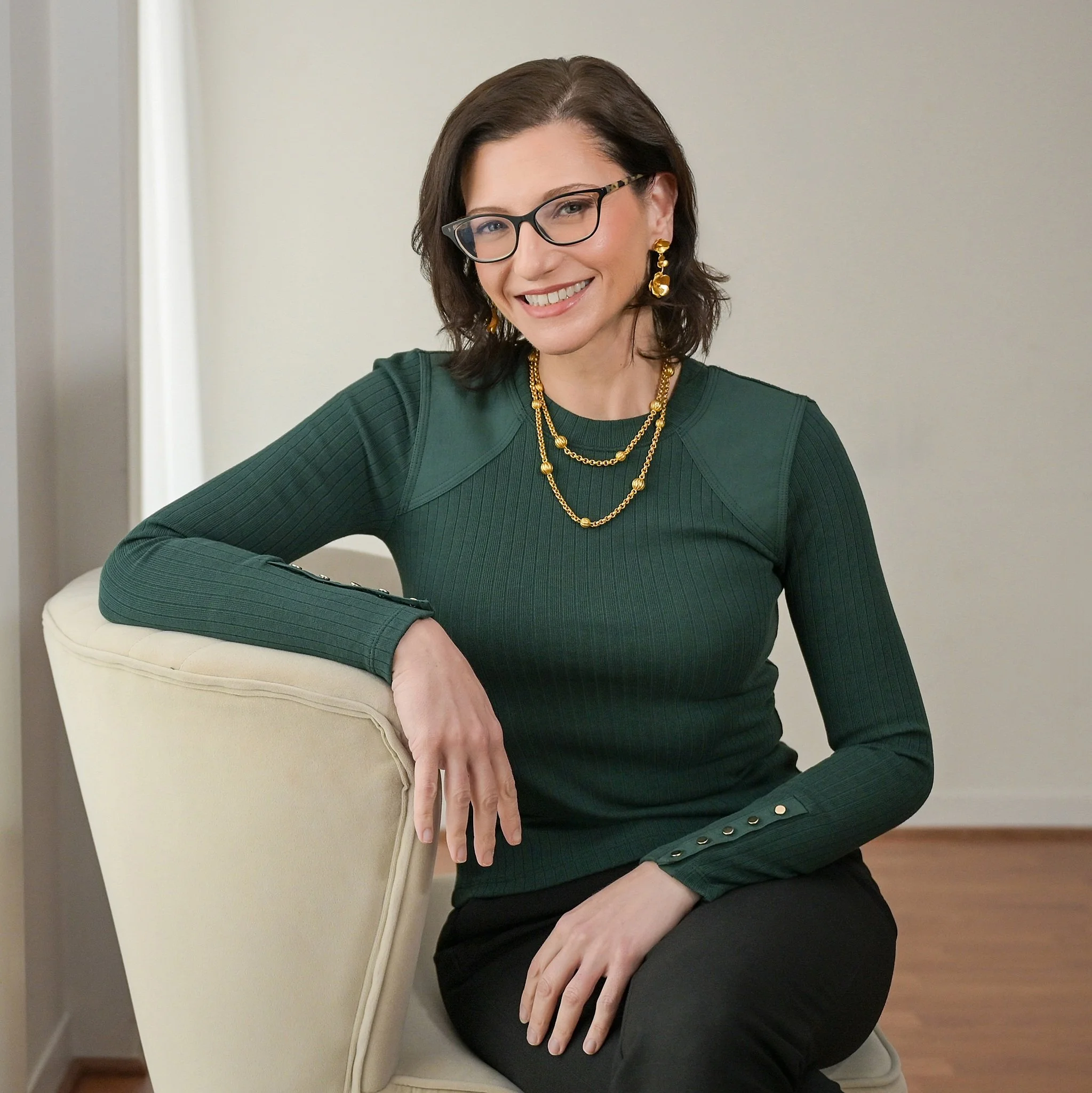 Woman with dark hair, glasses, and gold jewelry sitting on a cream-colored chair in a room with light gray walls and wooden floors.