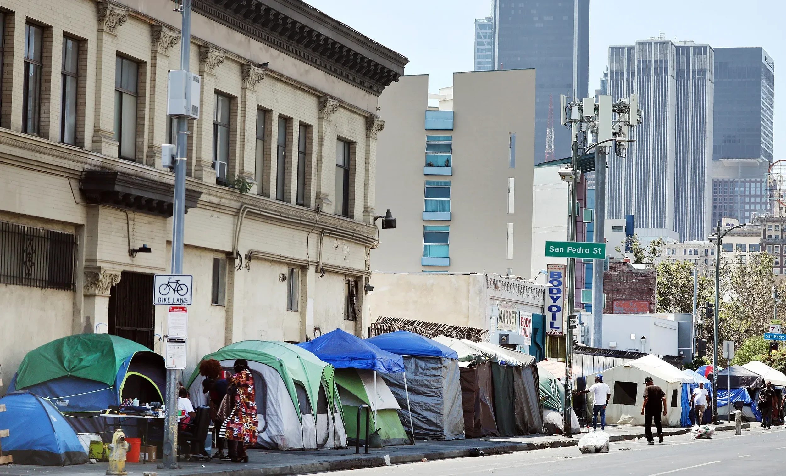 A city street scene with numerous tents set up along the sidewalk, people walking, and tall buildings in the background. There is street signage for San Pedro Street and various storefront signs.