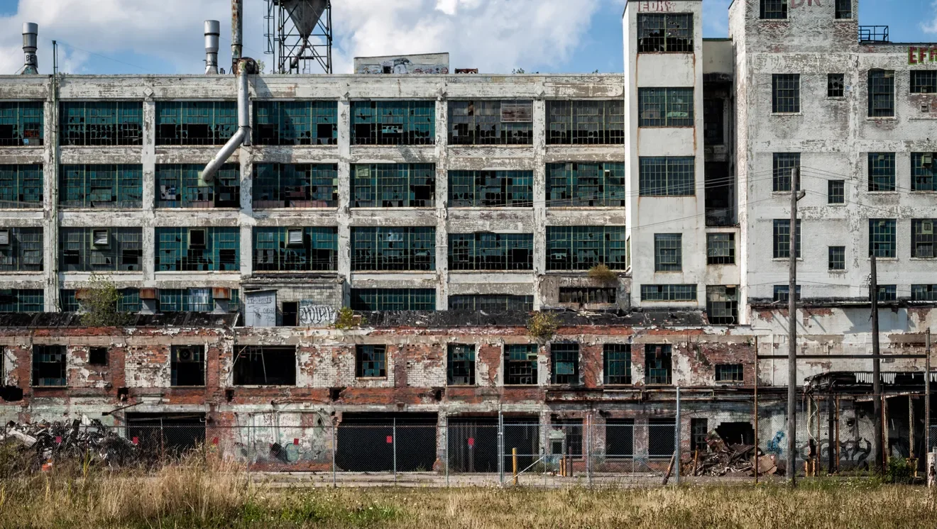 An abandoned, dilapidated building with broken windows, peeling paint, and overgrown grass in the foreground.