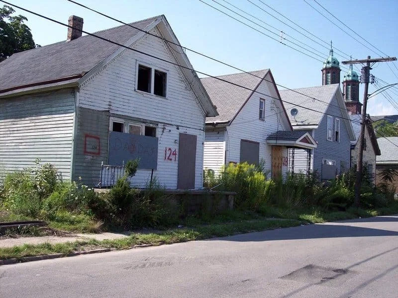 Old, abandoned white houses with boarded-up windows and overgrown yards on a quiet street.