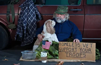 Older man with a beard sitting at a table with a dog, holding a sign that says 'Homeless Vet Support Your Troops'