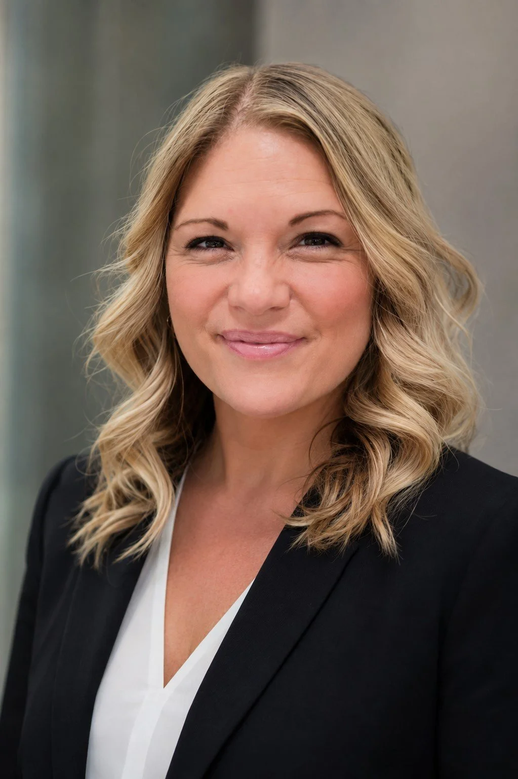 Portrait of a woman with wavy blonde hair, wearing a black blazer and a white blouse, smiling softly against a neutral background.