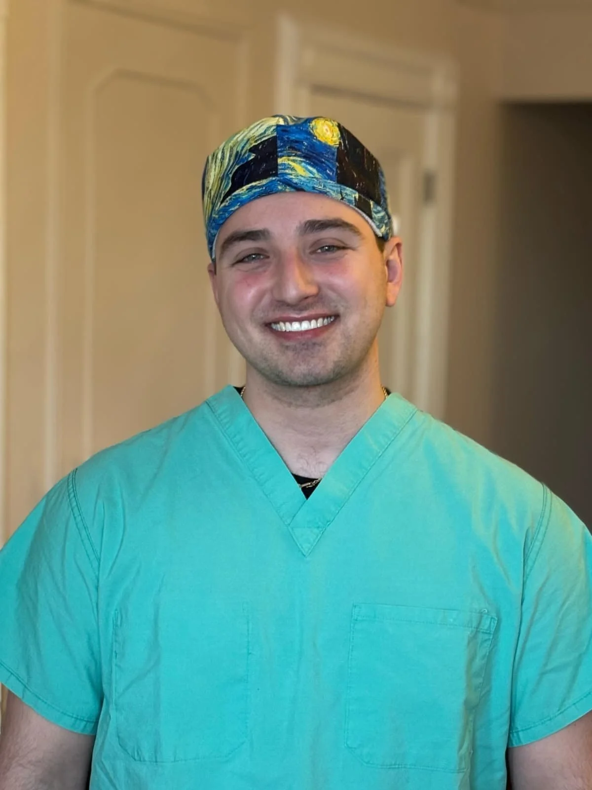 A smiling man in green medical scrubs and a colorful cap, standing inside a room with beige walls and white doors.