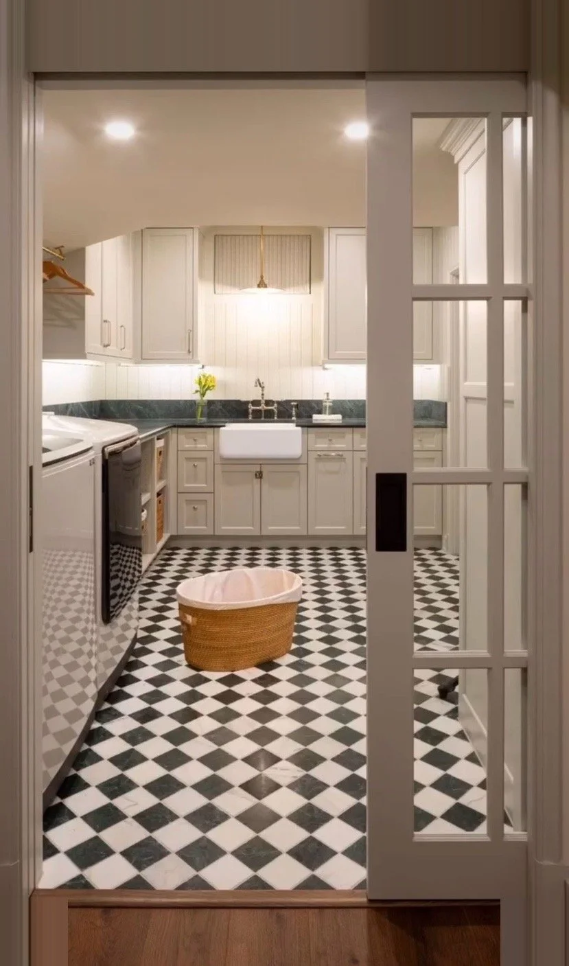 View through a glass-paneled door into a laundry room with black and white checkered floor. The room has white cabinets, a white farmhouse sink, a laundry machine, a small orange laundry basket, and a vase with yellow flowers on the countertop.