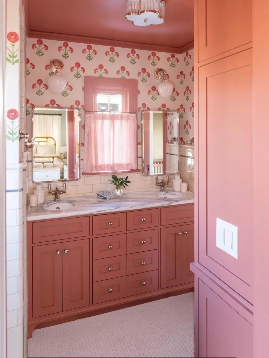 Bathroom with pink cabinetry, floral wallpaper, two mirrors, pink curtains, and vintage-style fixtures.