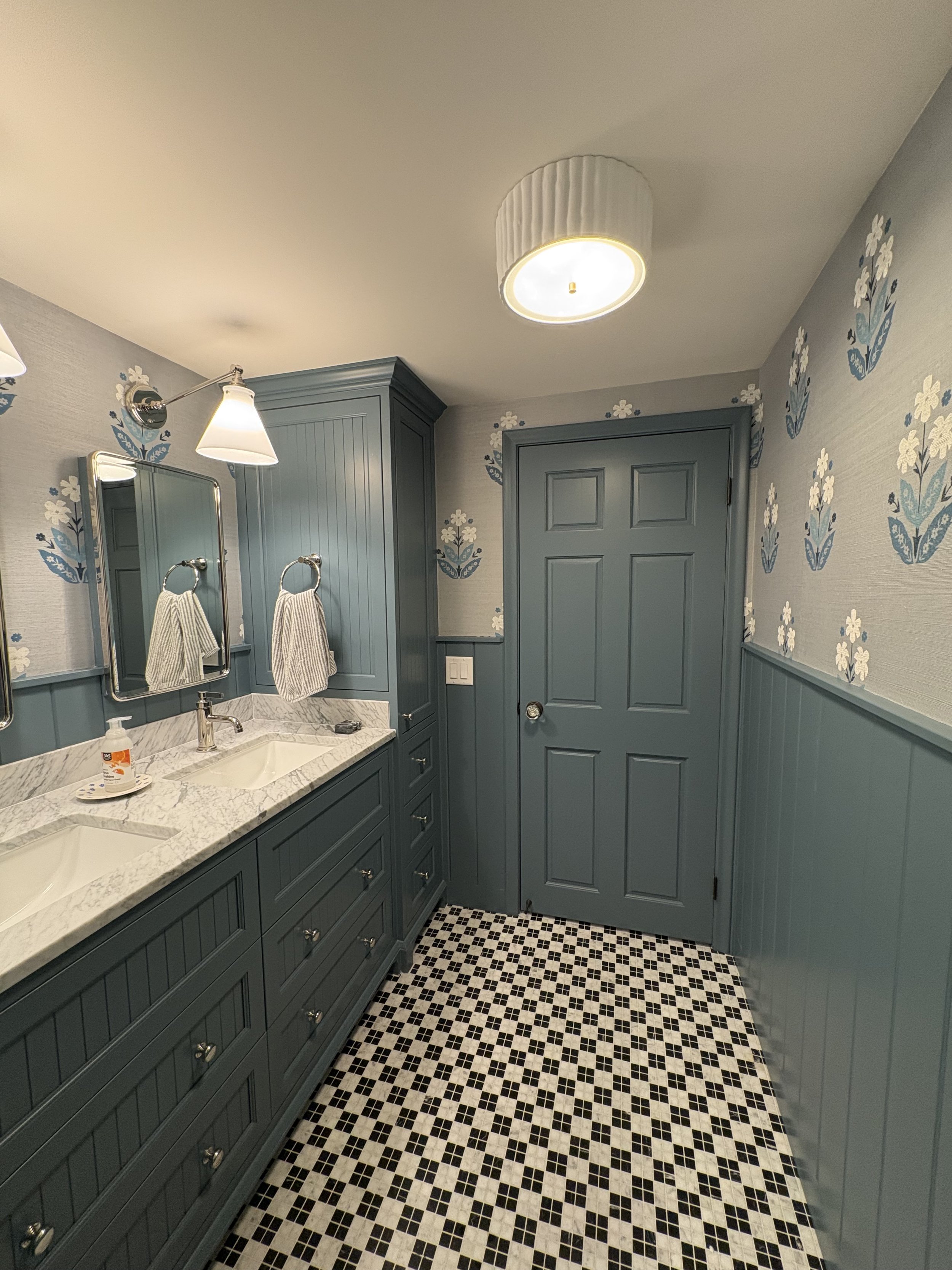 A bathroom with blue cabinets, a marble countertop, two mirrors above the sink, floral wallpaper with white and blue flowers, and a black and white tiled floor.