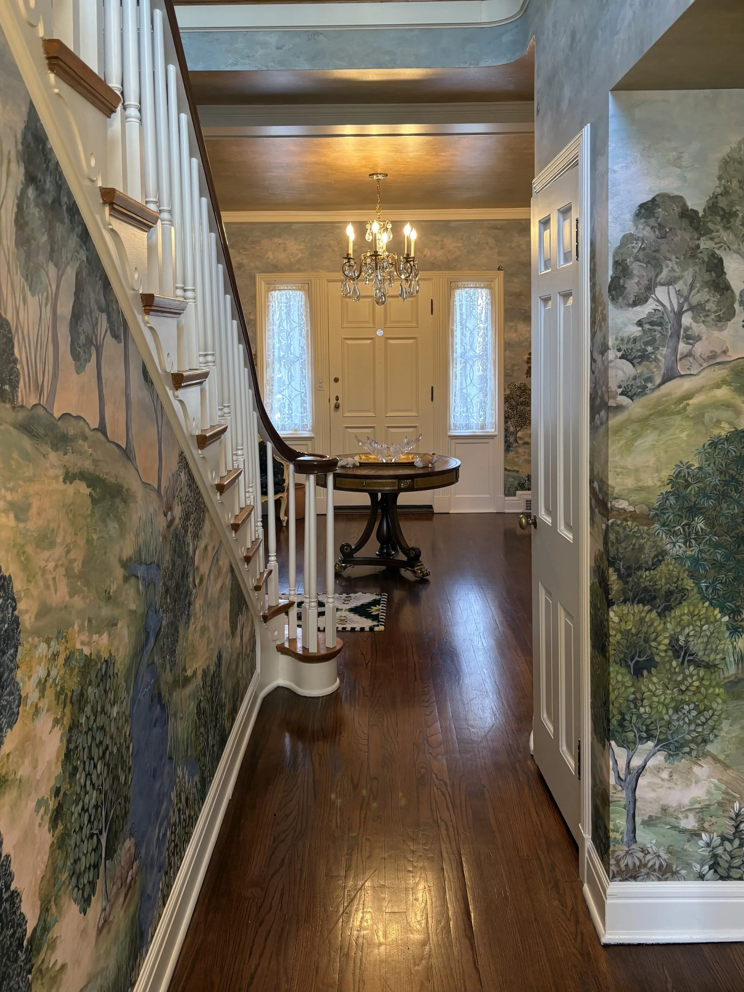 Entryway of a home with wooden floors, a chandelier, a front door, and painted murals of trees on the walls.