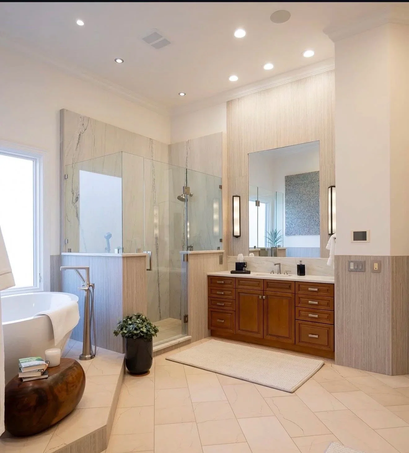 Modern bathroom with glass-enclosed shower, wooden vanity with mirror and lights, freestanding bathtub, window, potted plant, and beige tile flooring.