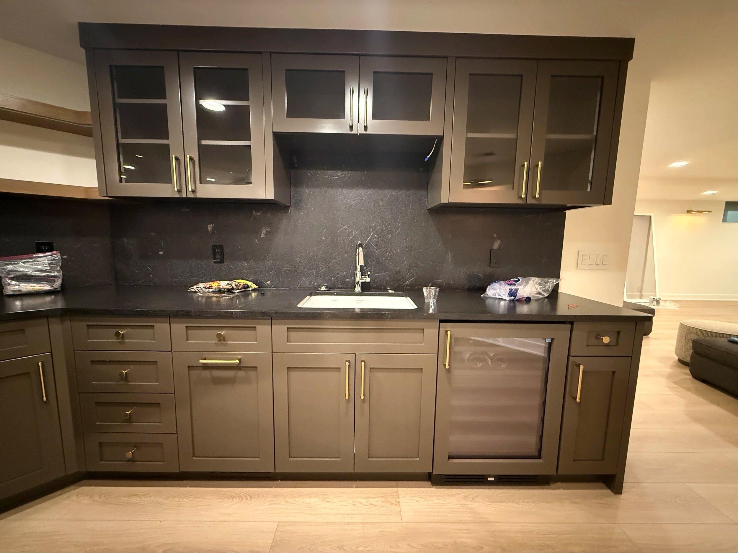 Gray kitchen cabinets with glass doors and gold handles, black countertop, and a black textured backsplash, with a sink and faucet, in a modern kitchen setting.