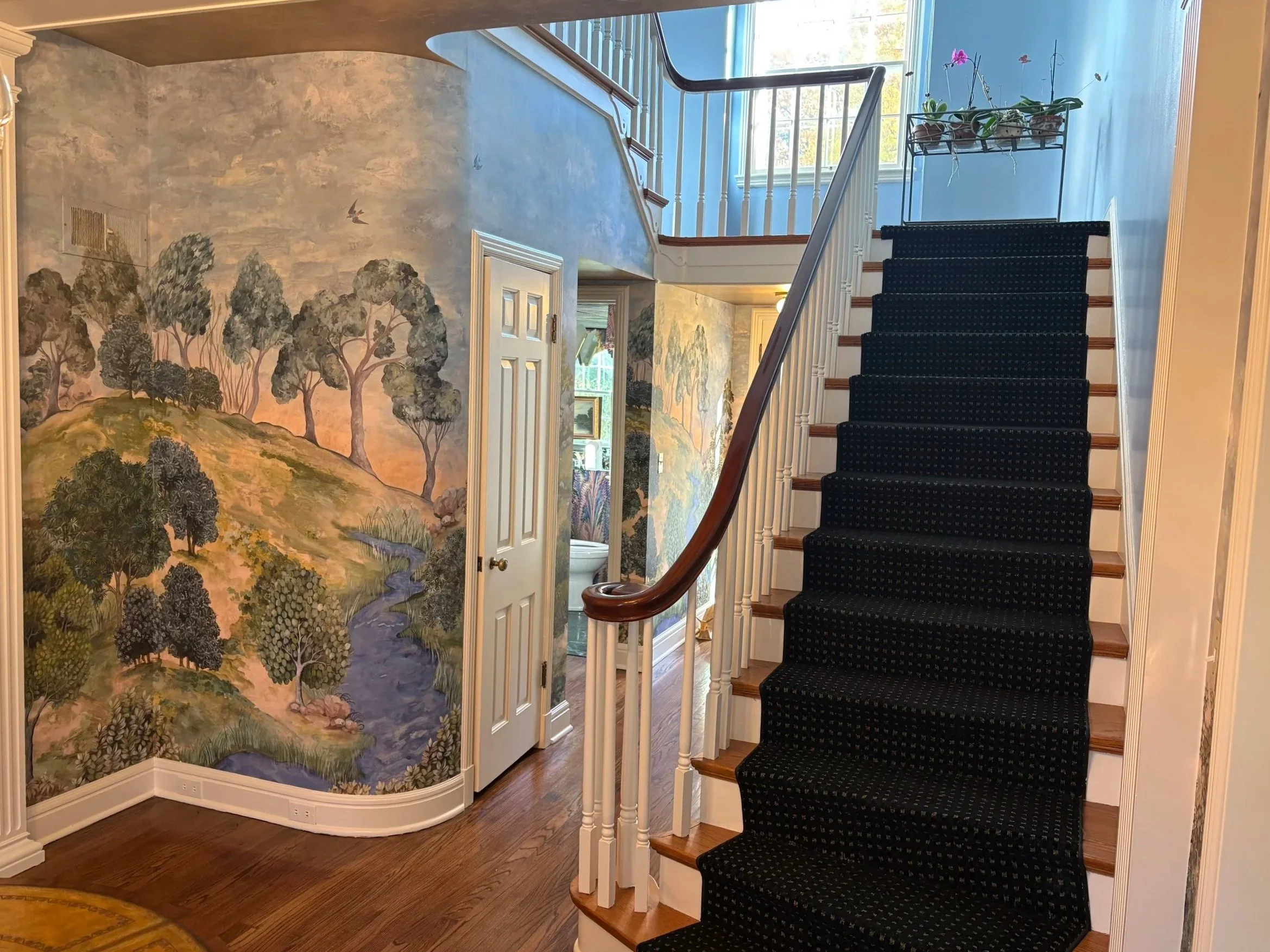 Interior view of a house staircase with a black polka dot runner, wooden steps, white balusters, and a curved handrail. A mural of trees and hills with a sky background is painted on the wall next to the staircase. Natural light streams through a win