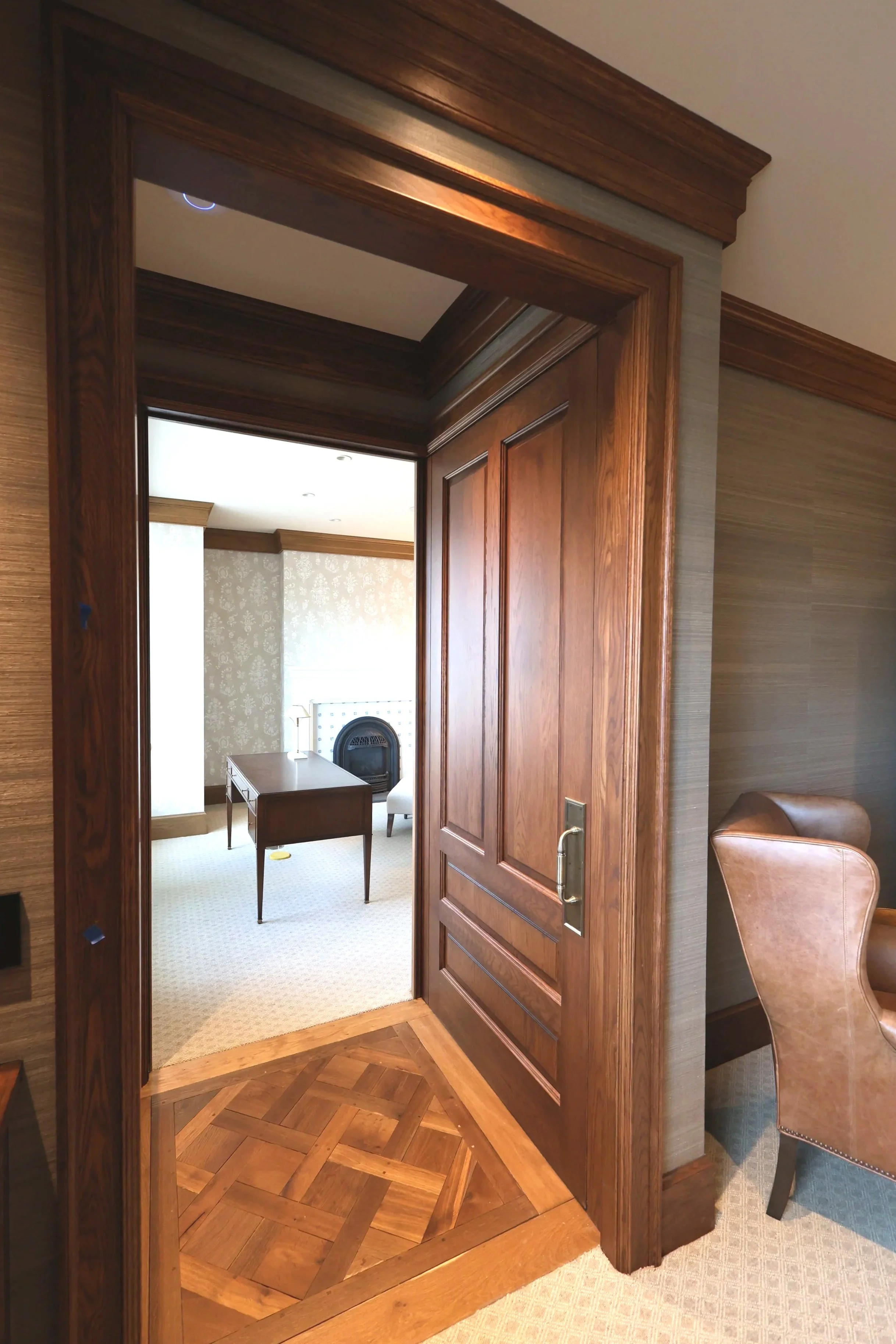 Wooden doorway with intricate parquet flooring leading into a room with a desk, a chair, and wallpapered walls. Part of a leather chair and a patterned carpet are visible.