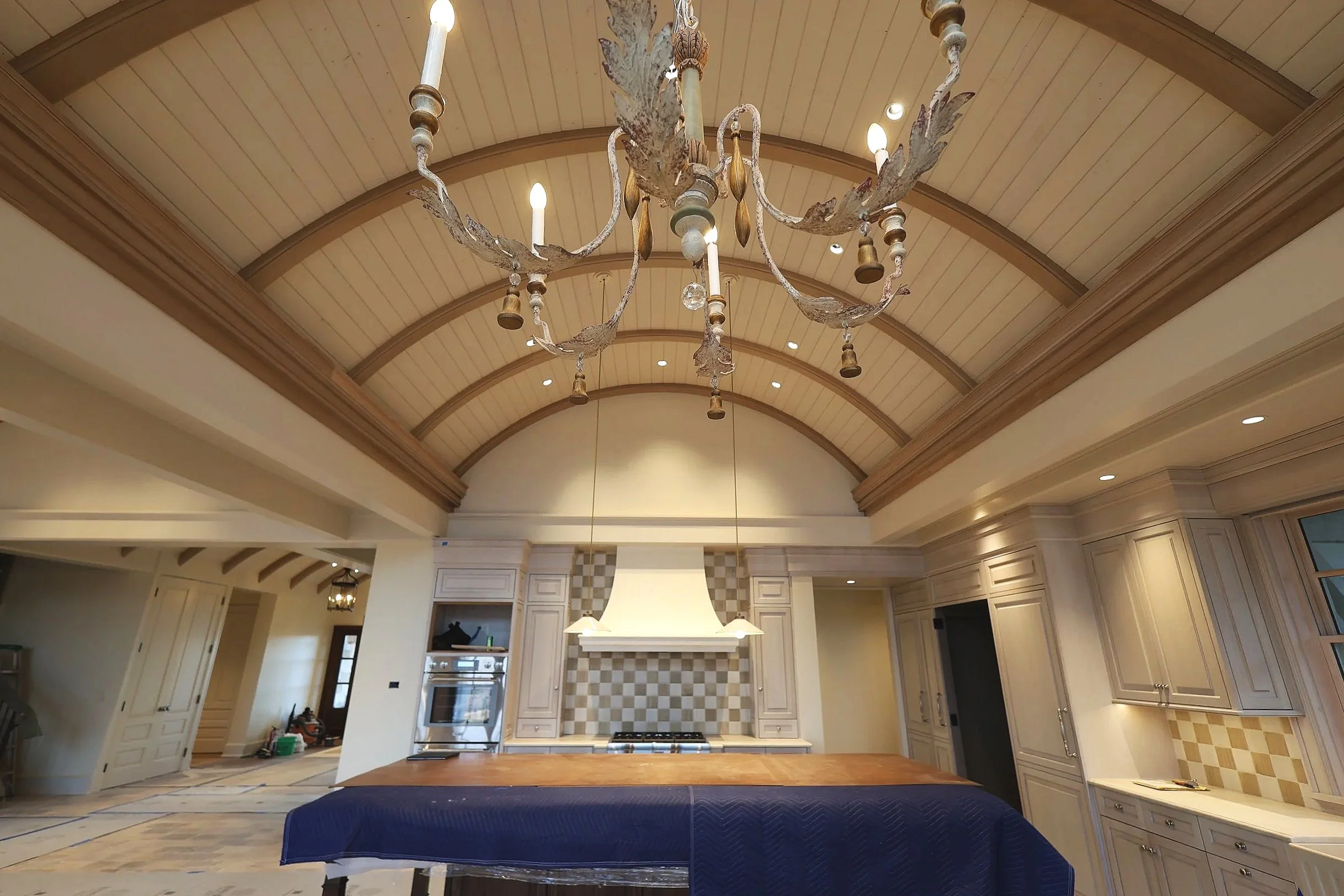 A chandelier with candle-shaped lights hanging from a wooden-beamed vaulted ceiling in a spacious kitchen with cream-colored cabinetry and tiled backsplash.