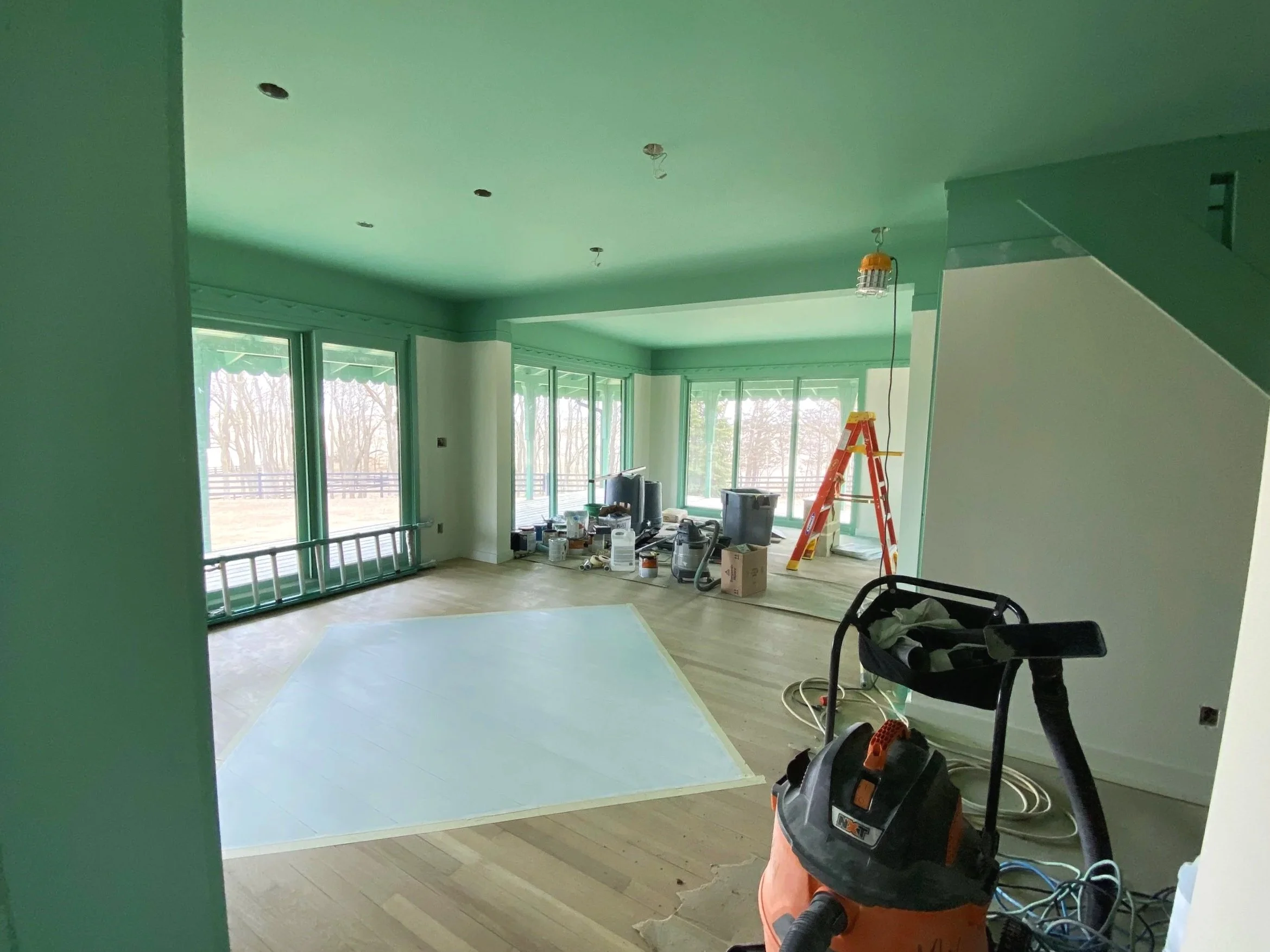 Living room under construction with paint supplies, a ladder, and a wet paint floor, large windows with a view of trees outside.