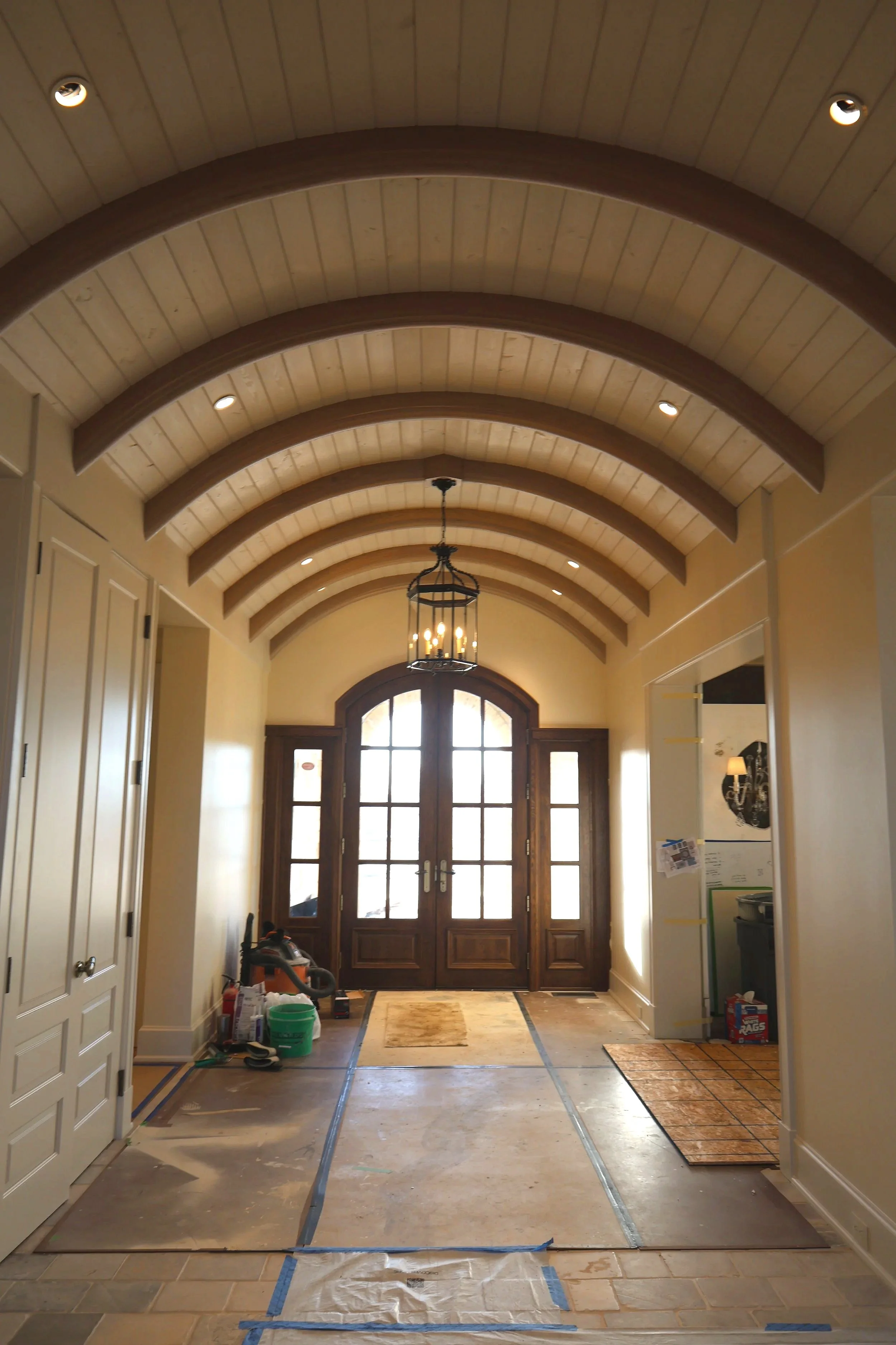 Entrance hallway of a house under renovation with a high arched wooden ceiling, an ornate chandelier, double front doors with glass panes, and partially completed flooring.