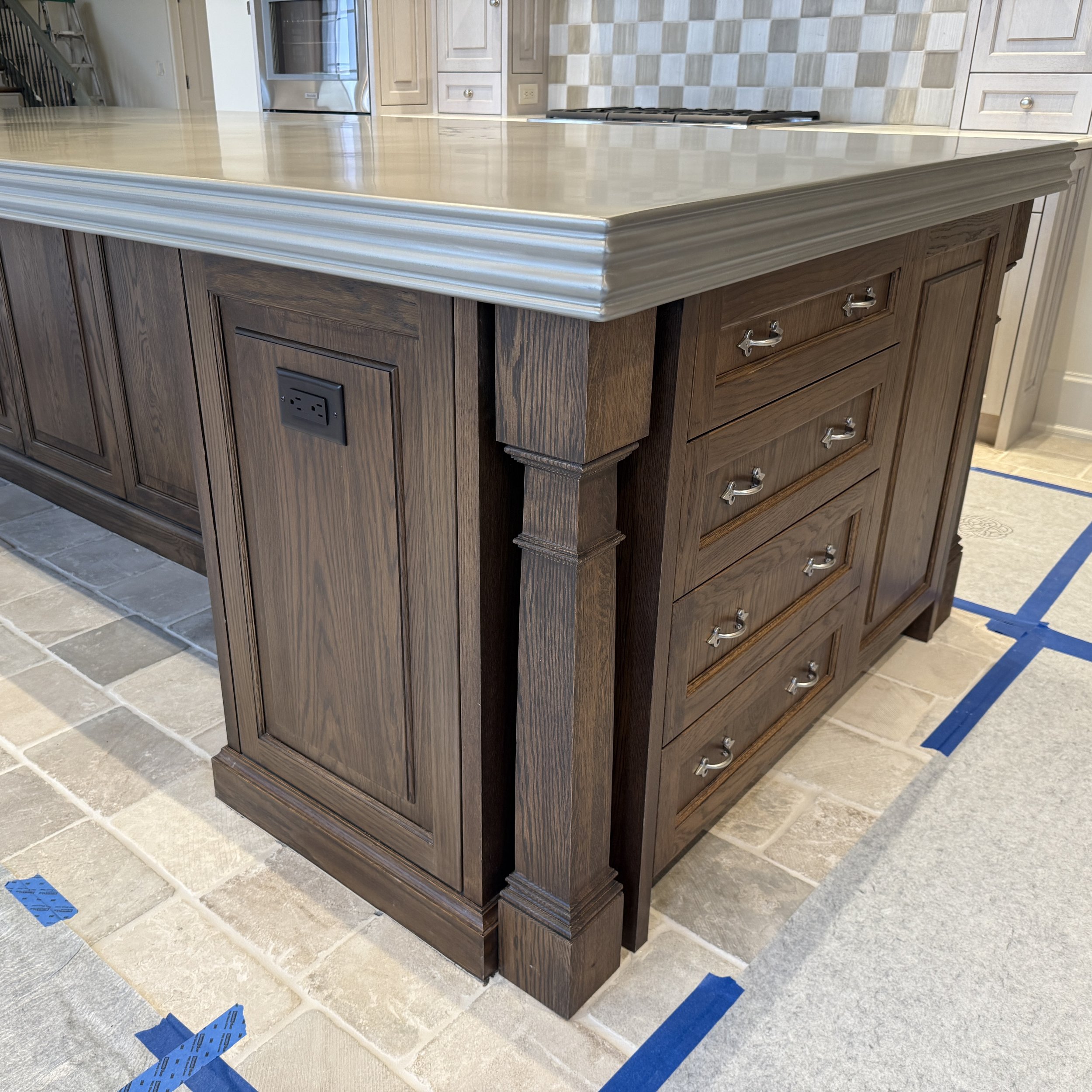A kitchen island with a beige marble top and a dark wooden base with drawers and cabinets. The floor has tiles with blue painter's tape in various places.