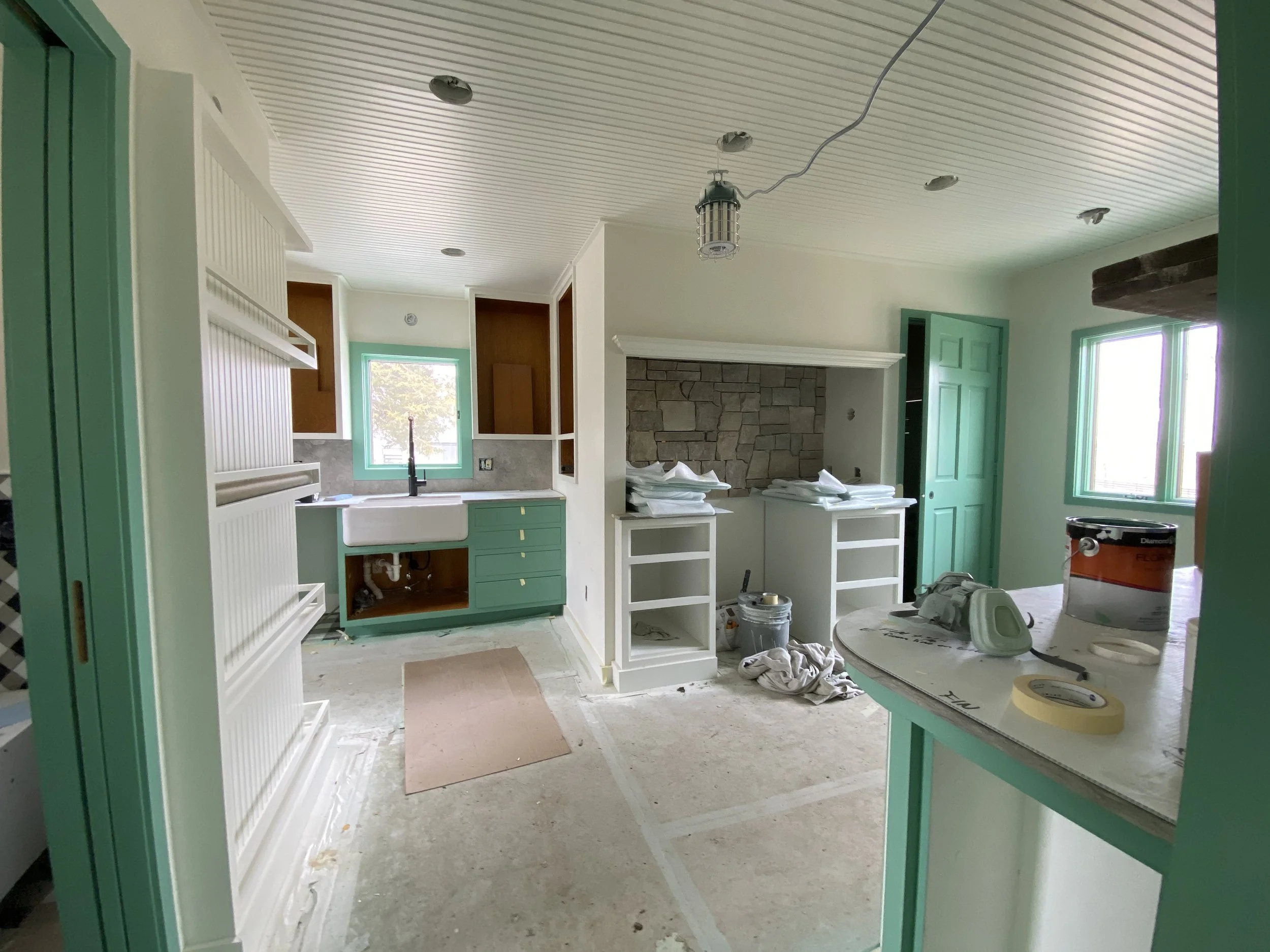 Interior of a kitchen under construction with green cabinetry, a white farmhouse sink, a stone fireplace, and ceiling lighting fixtures awaiting installation.