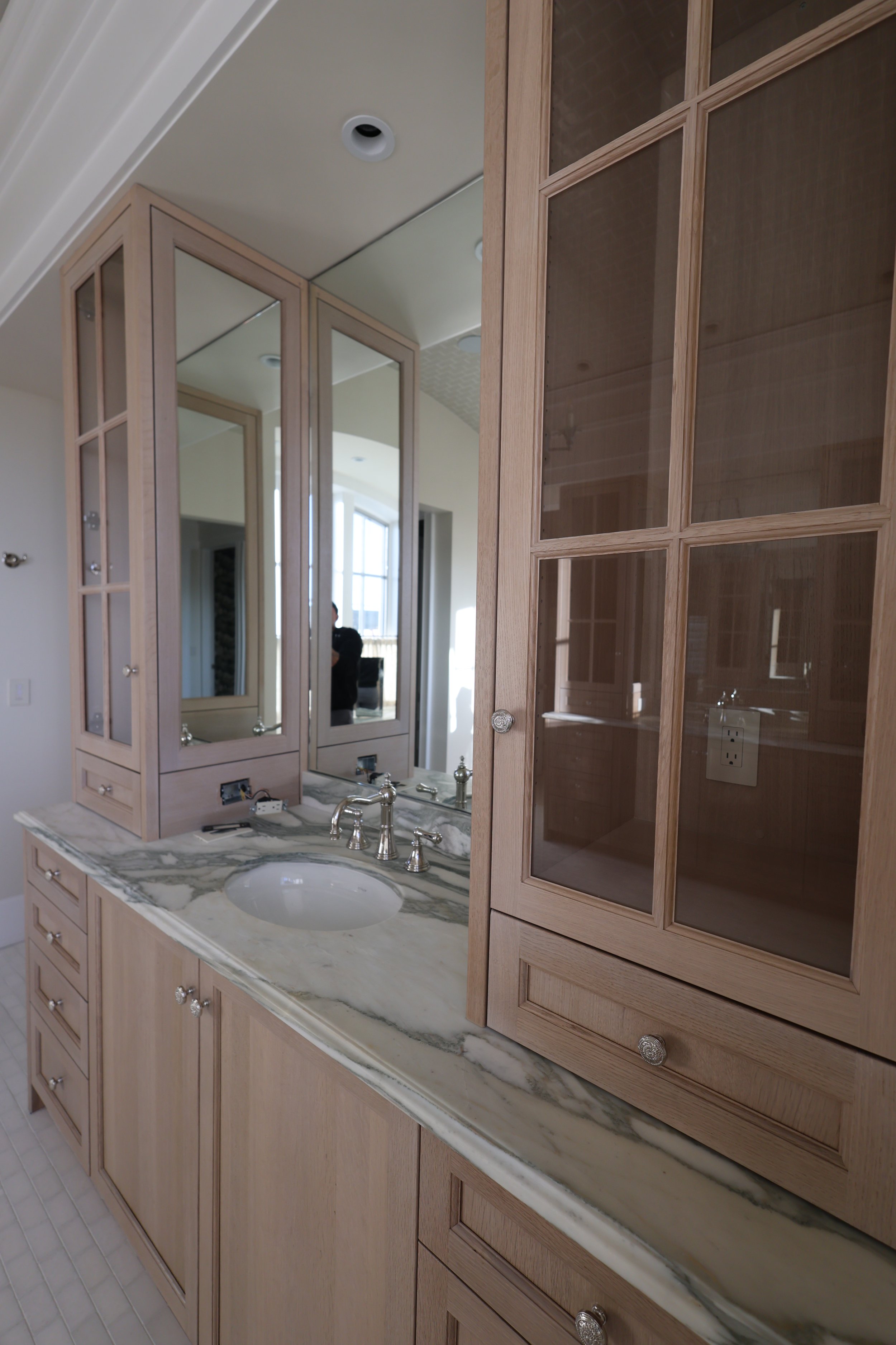 A bathroom vanity with a marble countertop, a undermount sink, a chrome faucet, and wooden cabinets with glass-paneled doors. A mirror and part of a window are visible in the background.