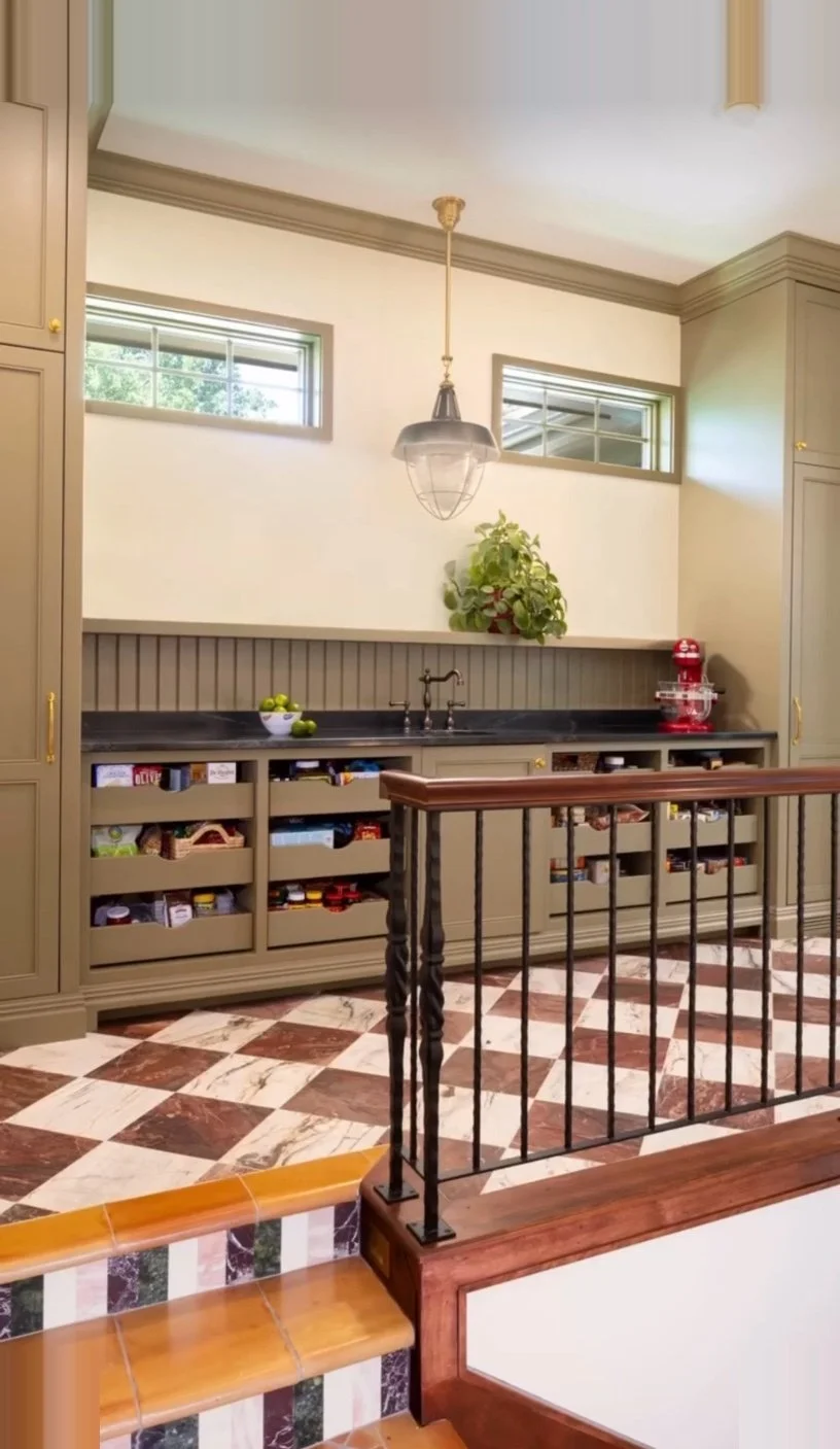 Kitchen with beige cabinets, black countertop, and a built-in snack storage. Features a tray of green apples, a small red appliance, a large plant, and a hanging light fixture. The floor has a checkered pattern of brown and white tiles, and the image
