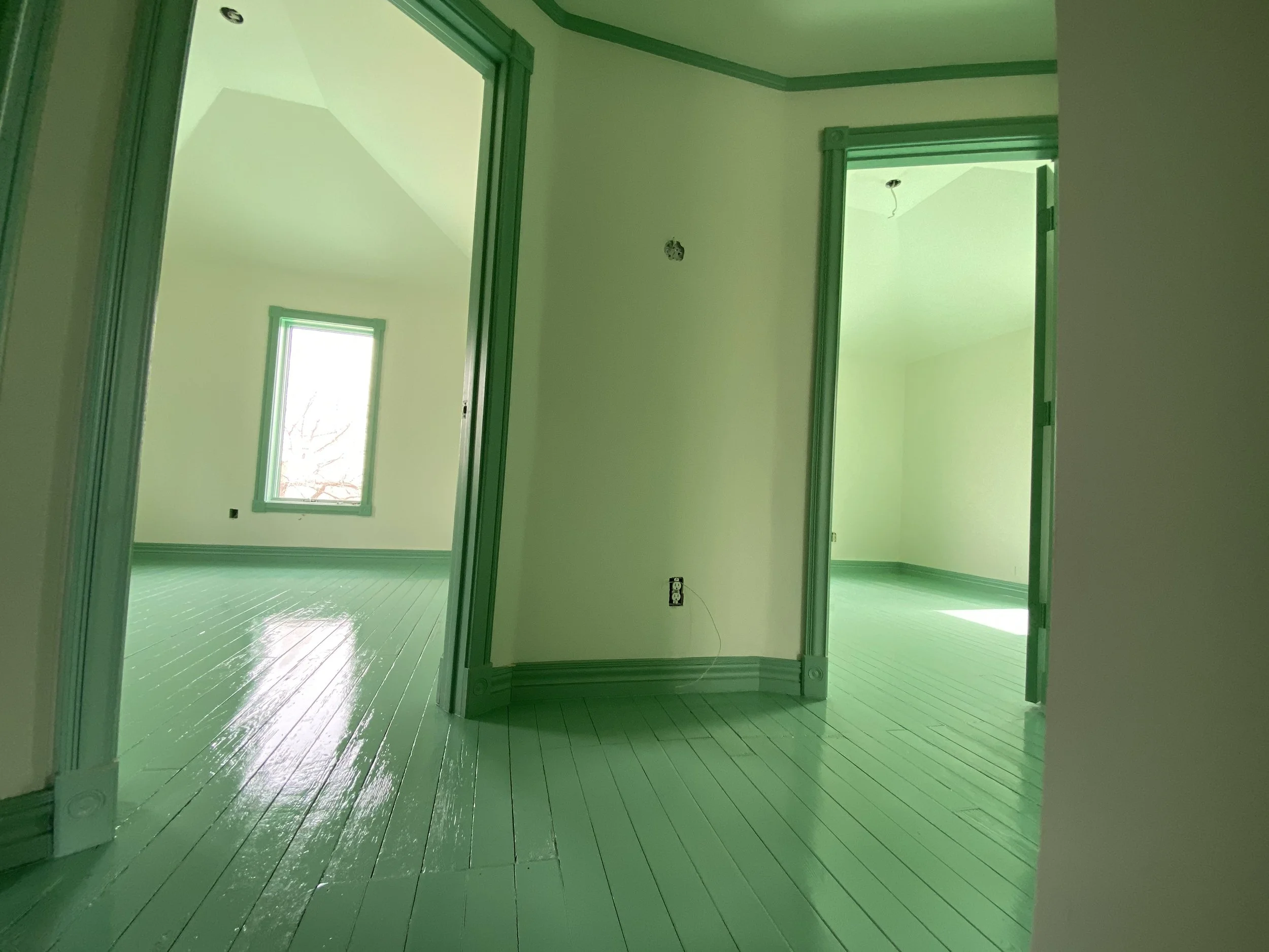 An interior room with freshly painted green walls, white trim, and a shiny green-painted wooden floor. There are multiple doorways and a window, all framed with green trim. The ceiling is high, and there's an electrical outlet and cable in the room.