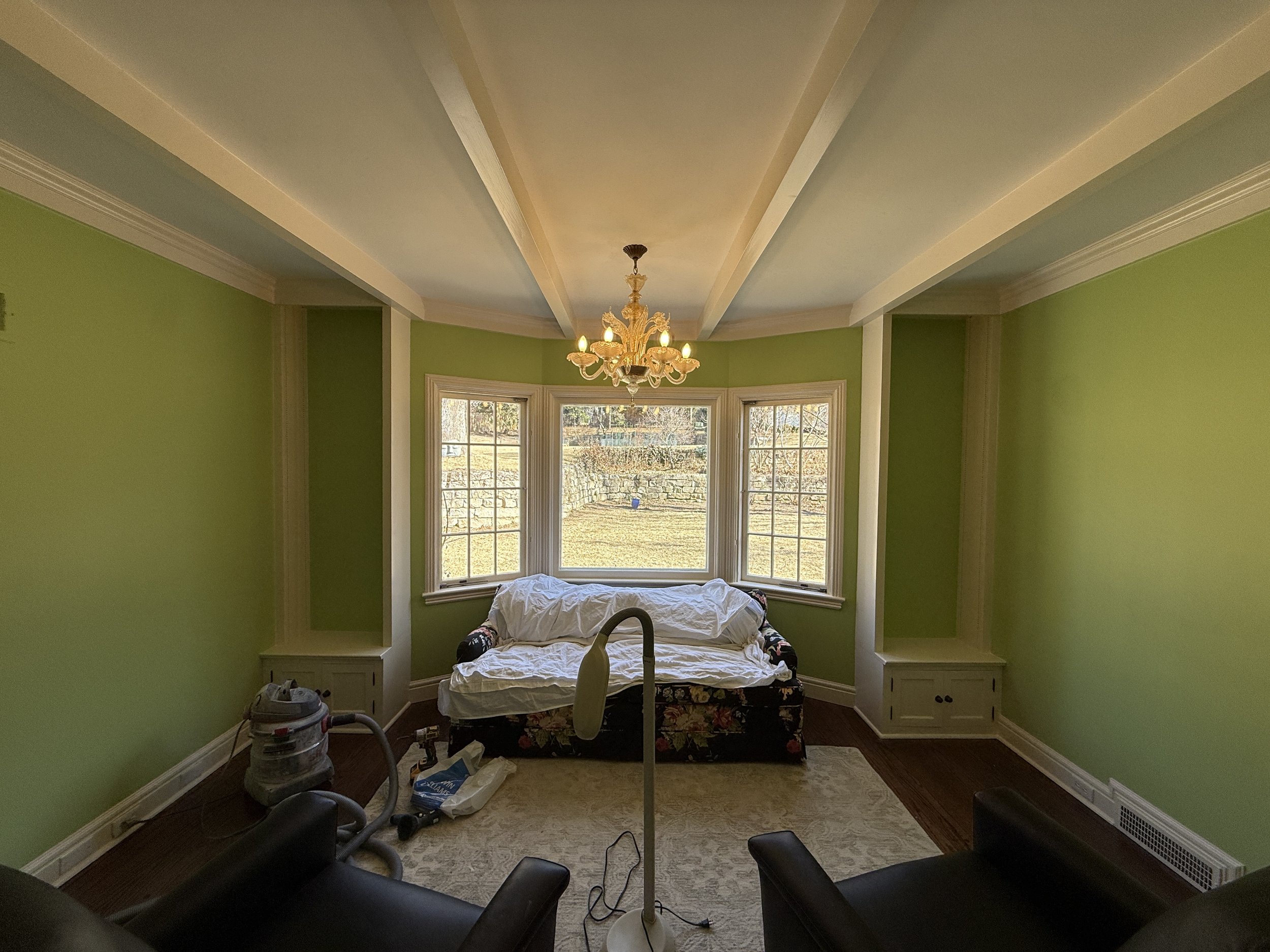 A room with green walls and large window, featuring a chandelier, a couch covered with a white sheet, and some cleaning equipment in the foreground.
