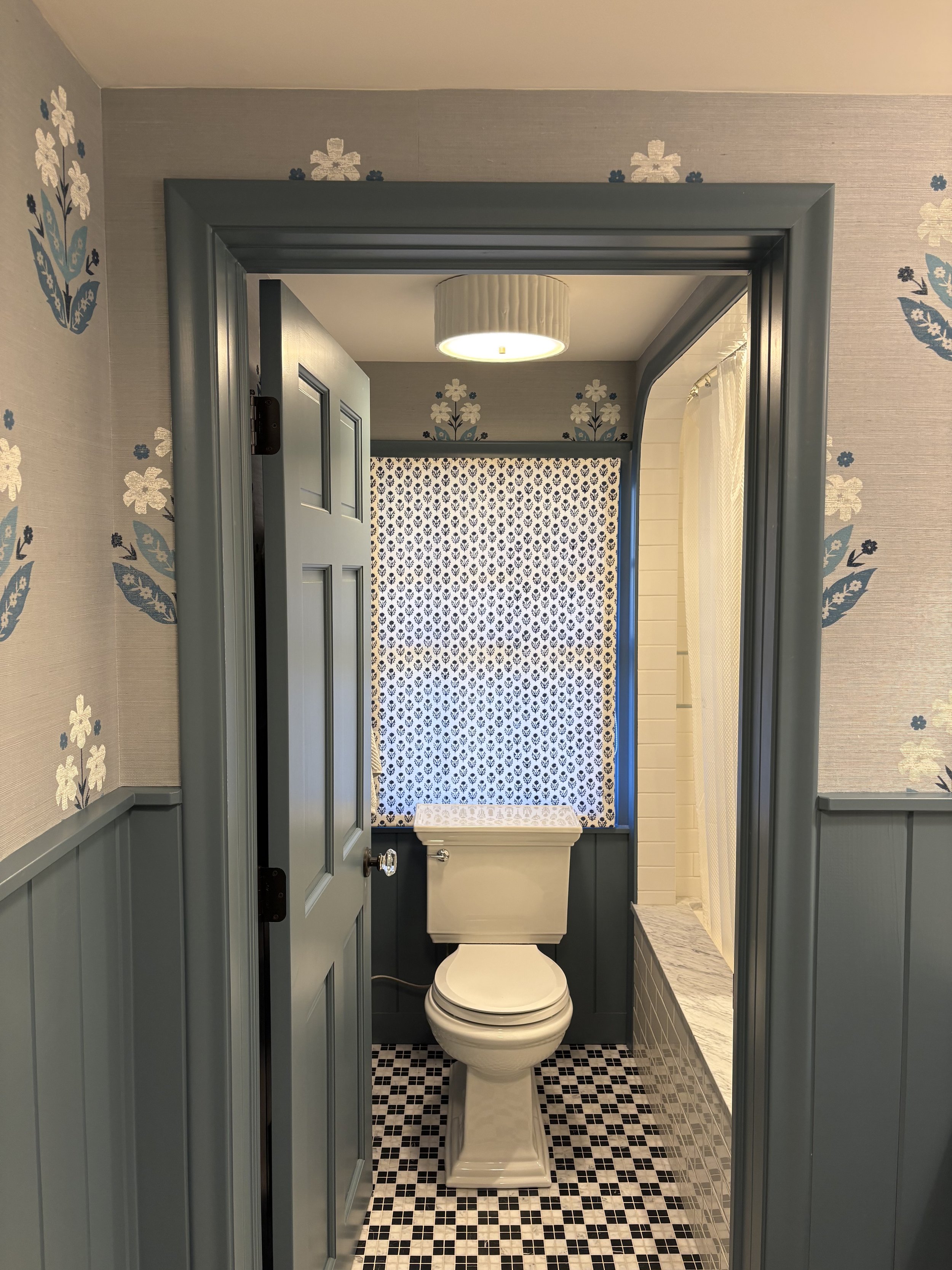 A small bathroom with a toilet in the center, surrounded by patterned wallpaper and a window with curtain, featuring a combination of blue and white decor.