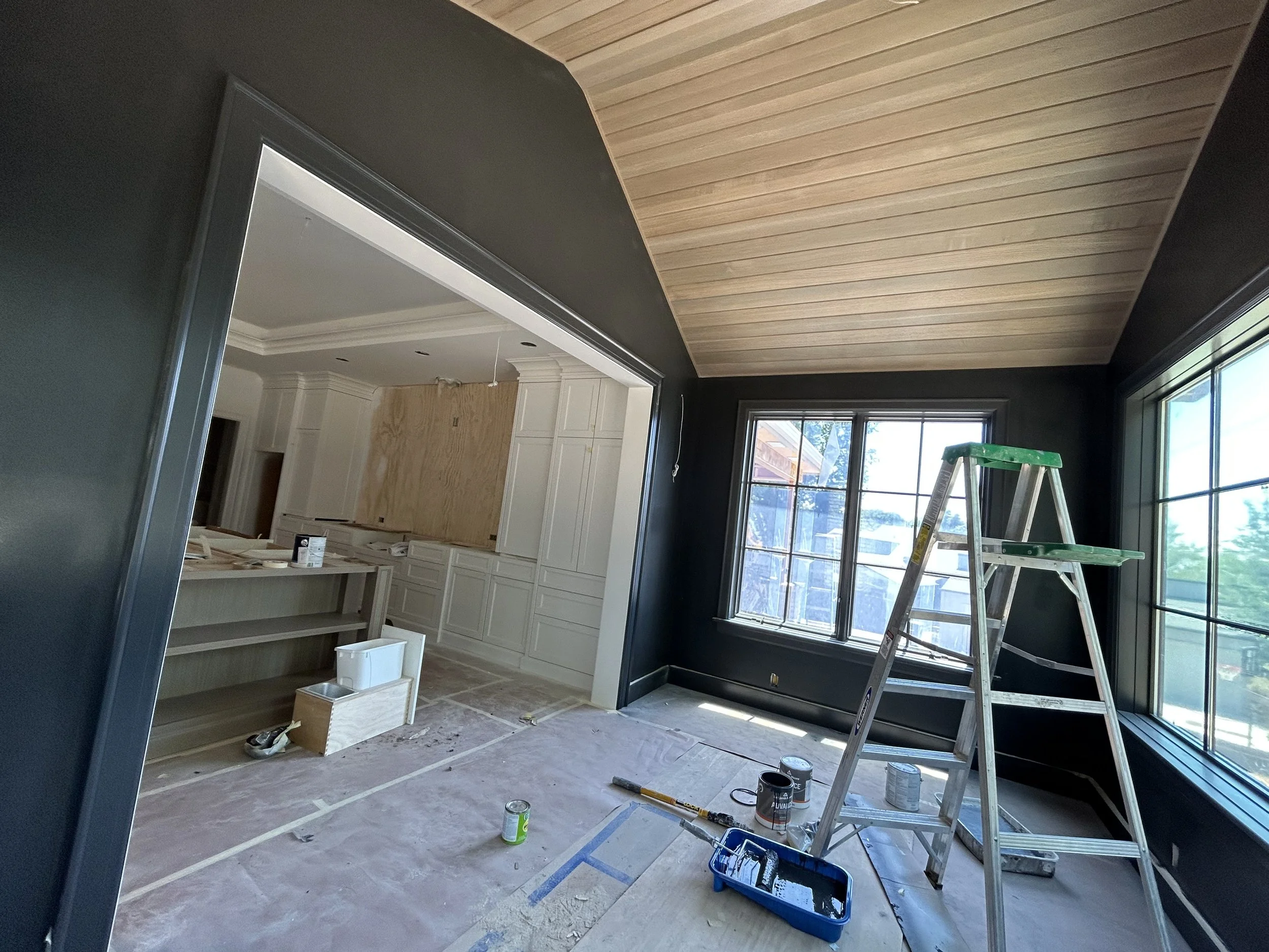 Interior view of a room under renovation, with a ladder, paint cans, and a partially painted ceiling and walls, large windows, and construction supplies on the floor.